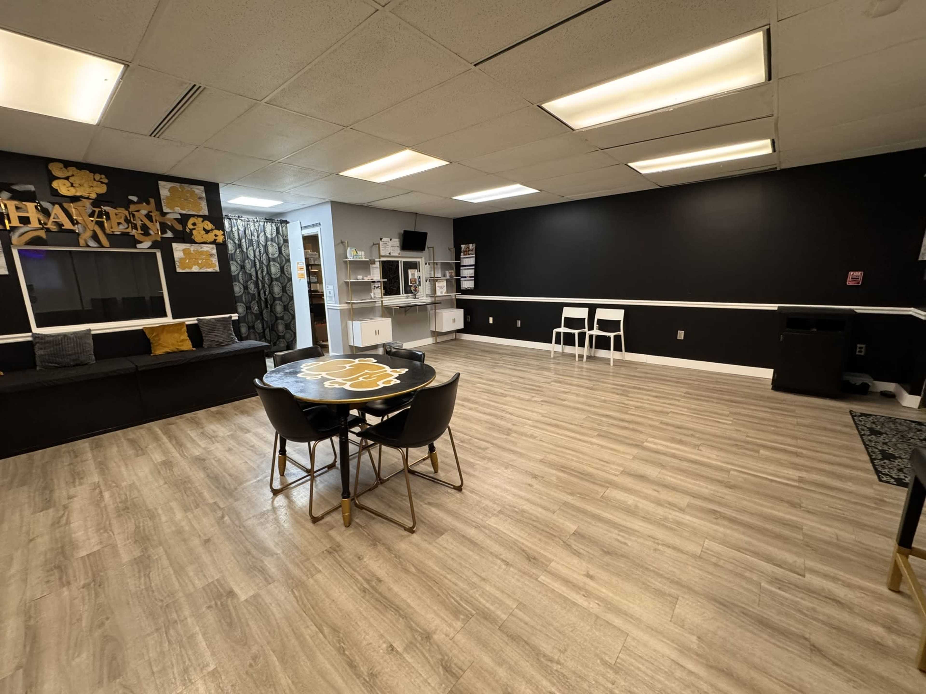 The image shows a spacious, well-lit room with a dark accent wall, a seating area with a sofa, a round table with chairs, and storage cabinets.