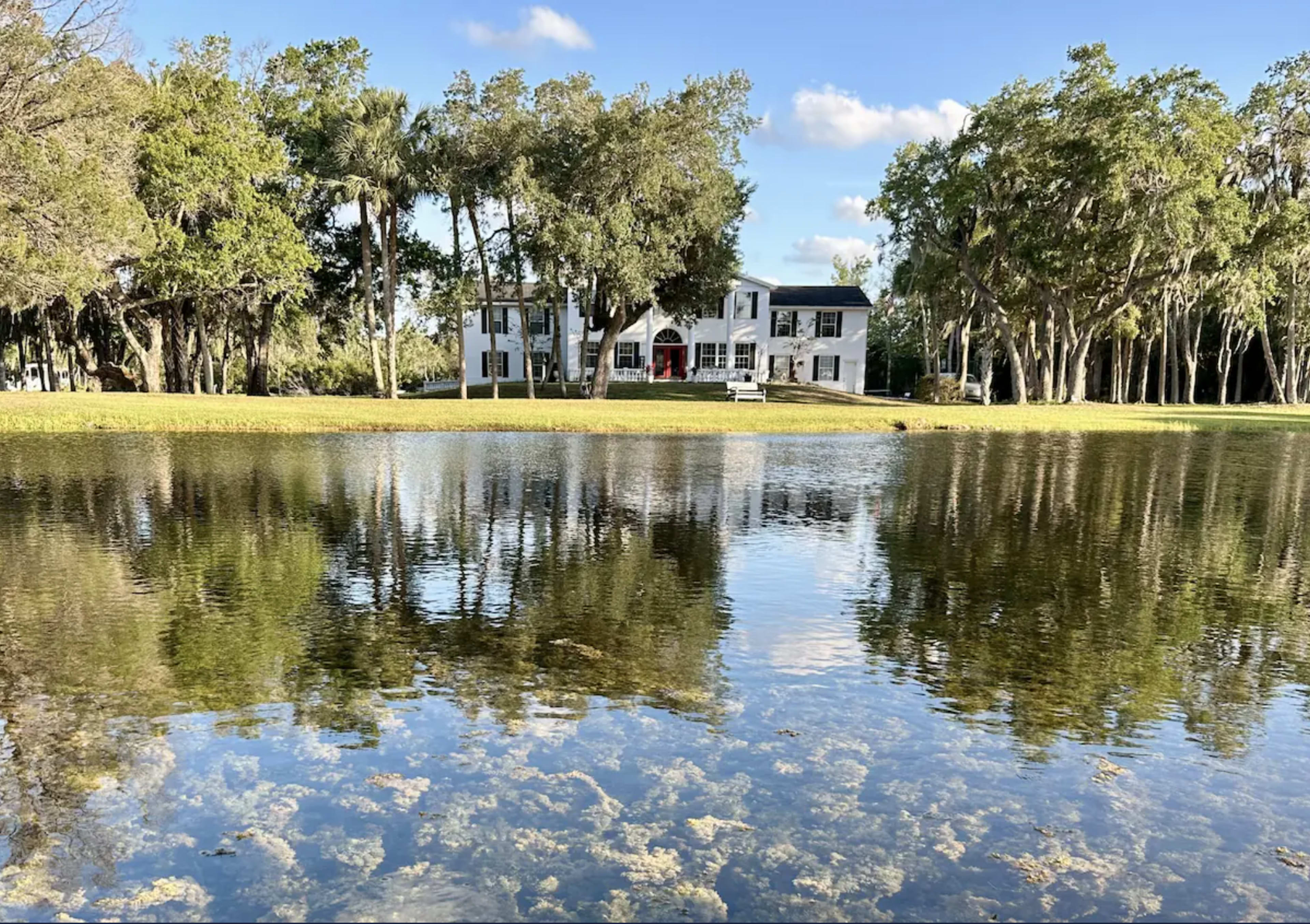 A white house with large windows stands by a clear pond surrounded by trees and grass.