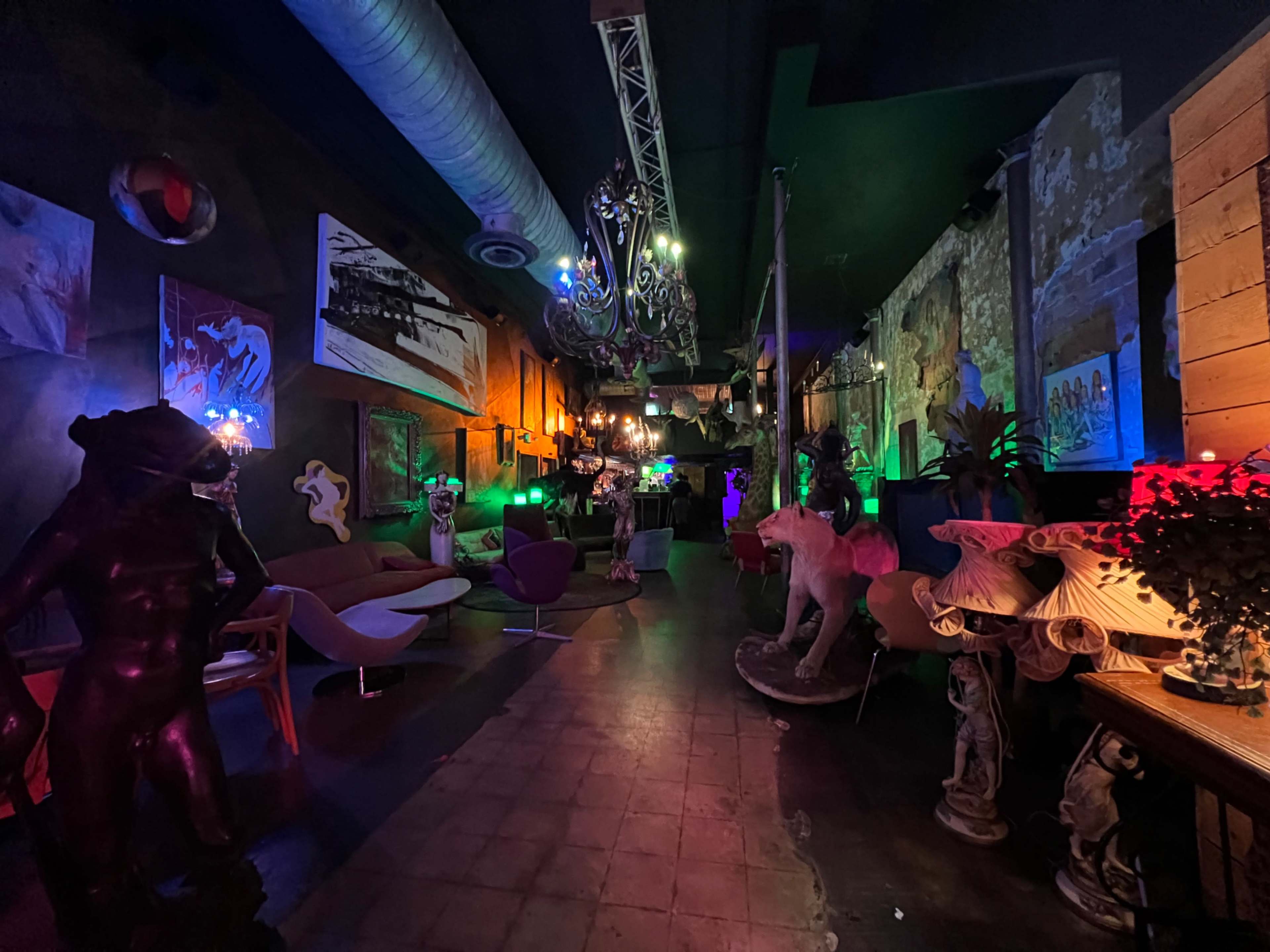 The image shows a dimly lit corridor in a vibrant art space, featuring sculptures, colorful lighting, and various artworks on the walls.