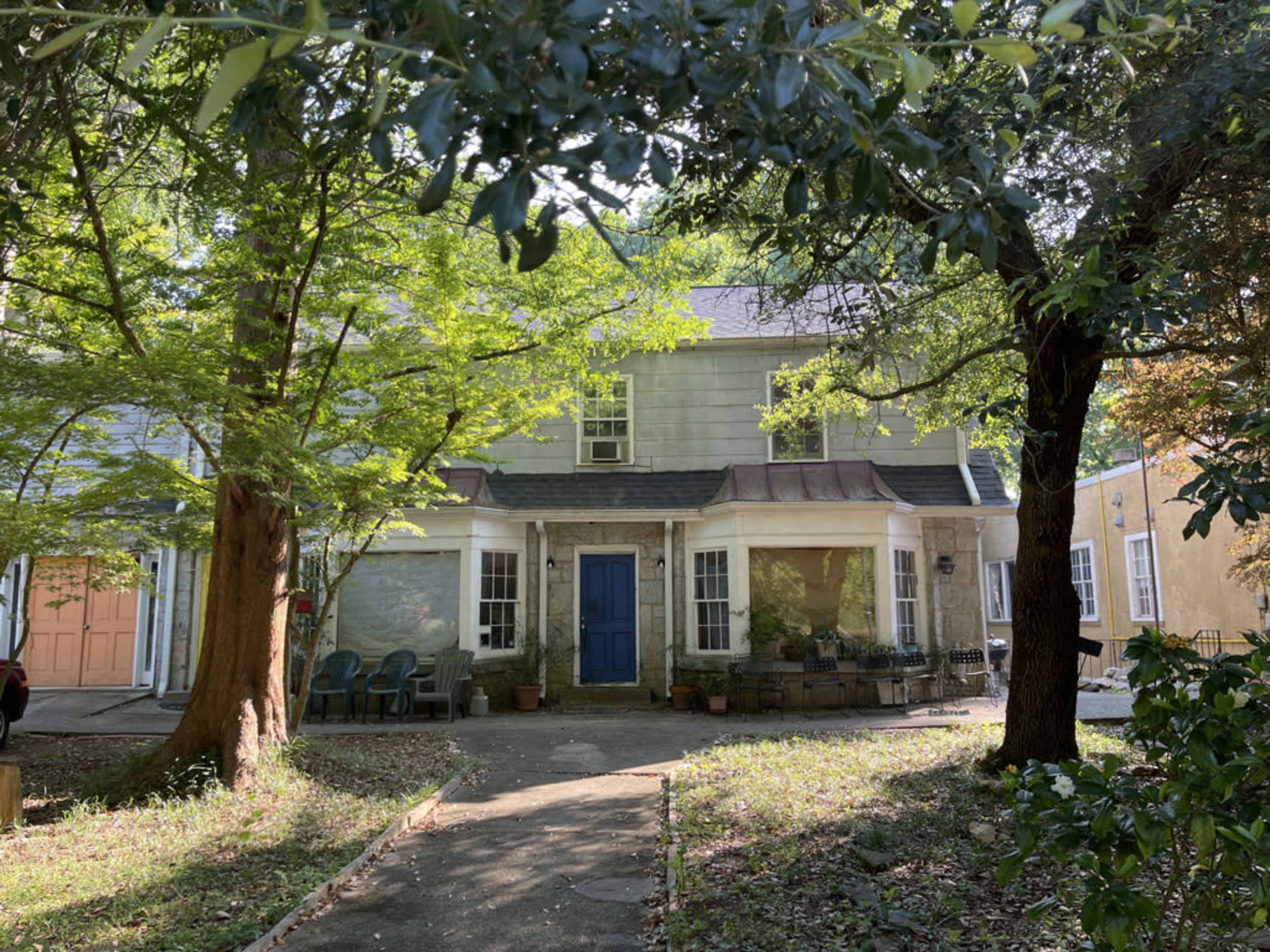 A two-story house with a blue front door is partially obscured by trees and surrounded by greenery.