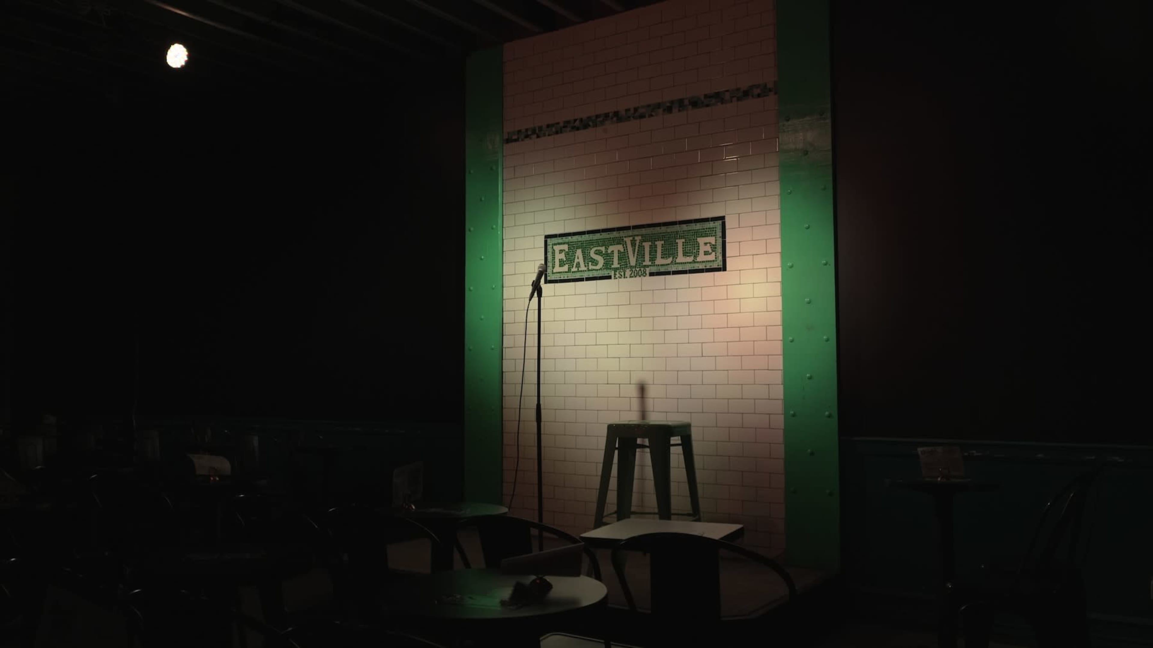 The image shows a spotlight illuminating a microphone stand beside a green stool in front of a tiled wall with "EastVille" written on it.