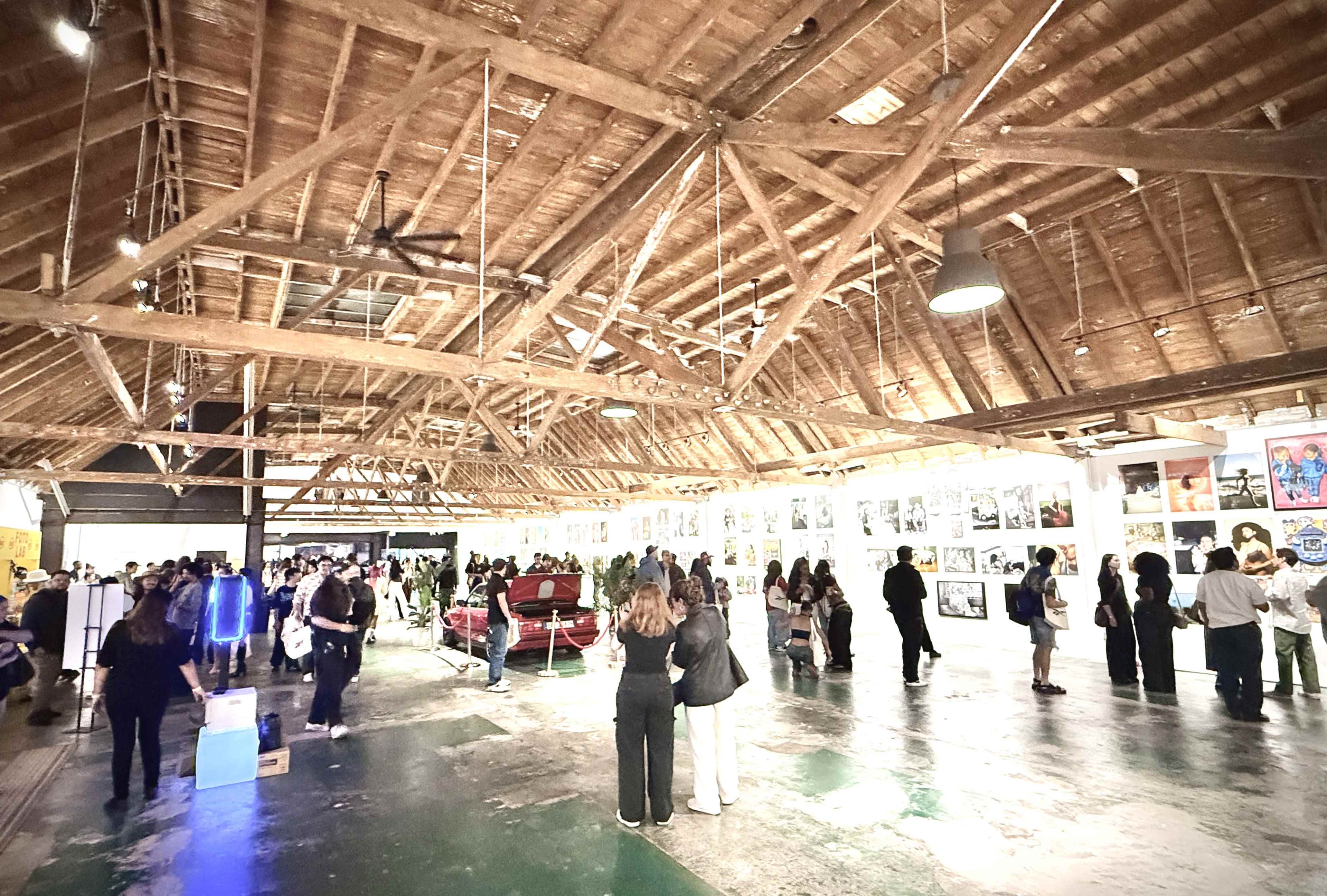 A large indoor gallery features a high wooden ceiling and is filled with people viewing numerous artworks displayed along the walls.