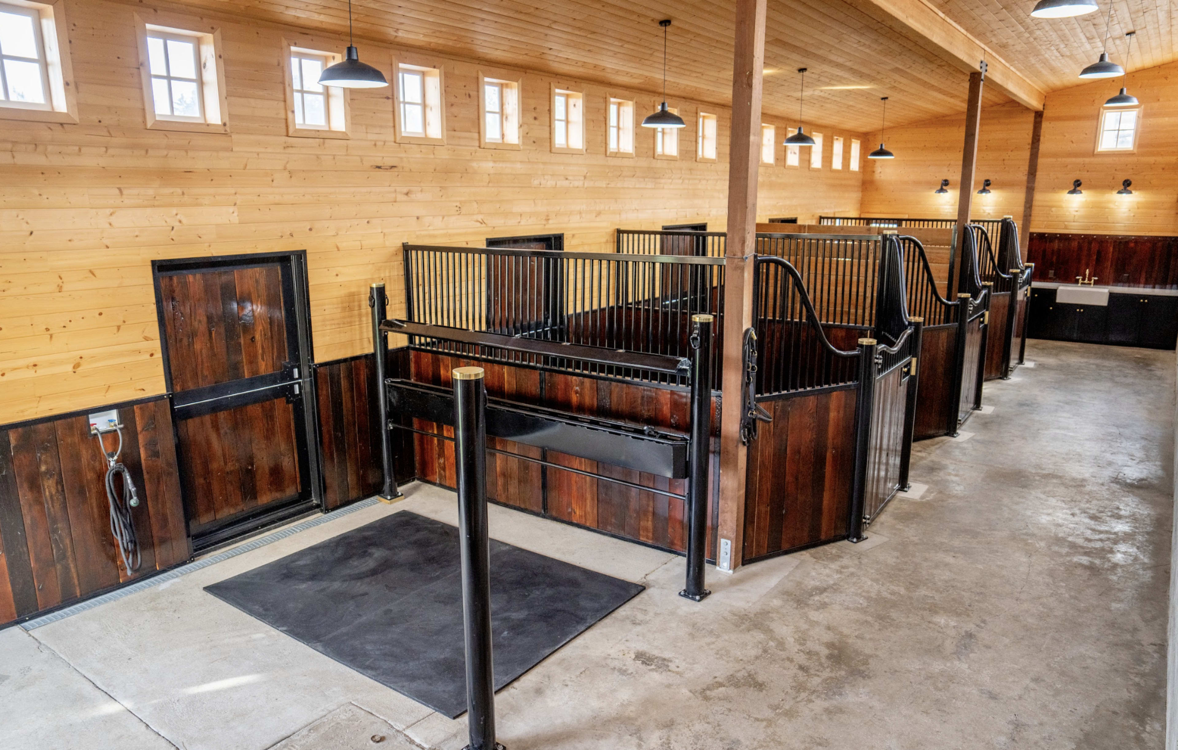 The image shows a spacious stable interior with wooden walls, several horse stalls on each side, and a concrete floor.