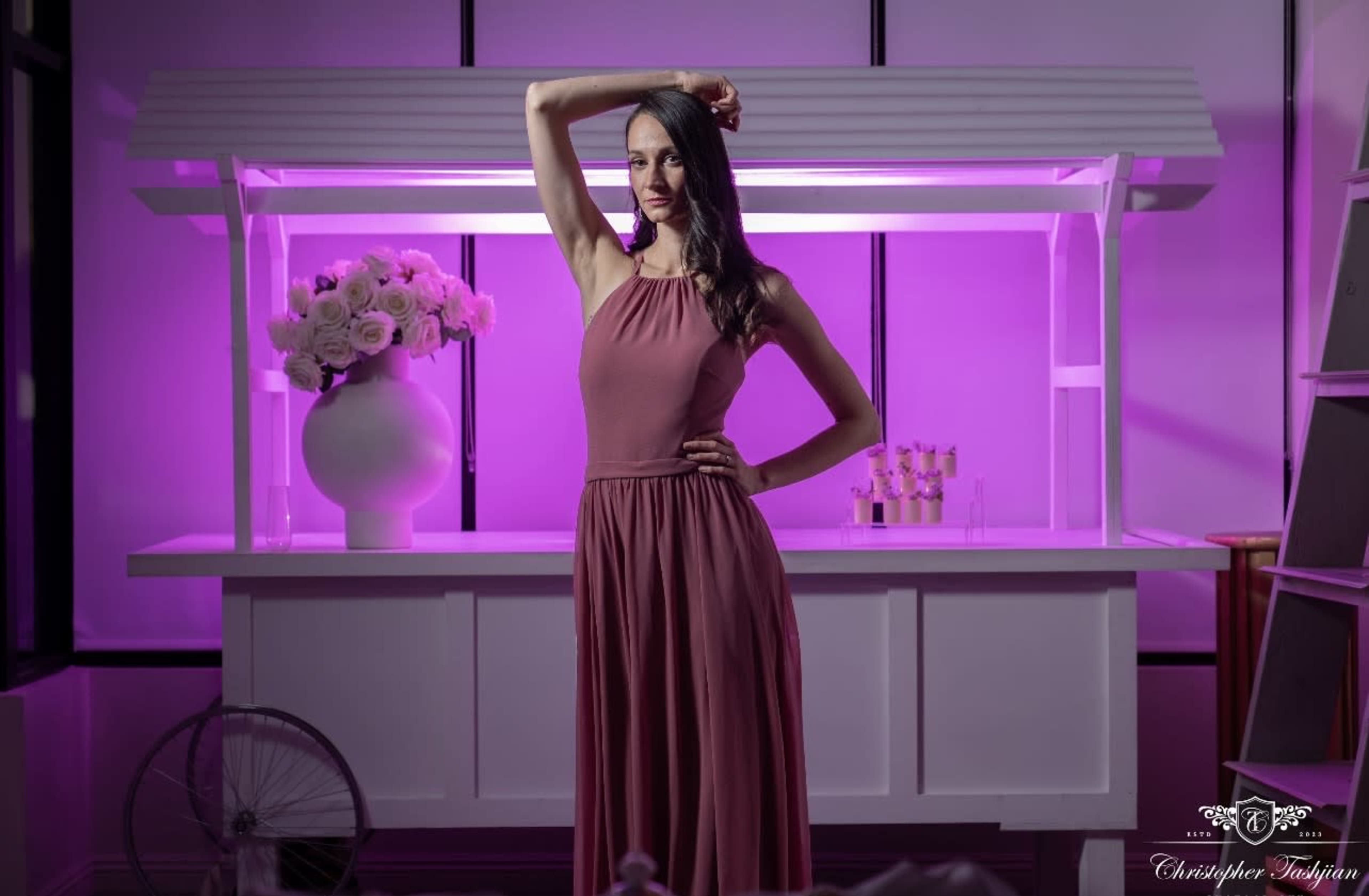 A woman in a pink dress poses confidently in front of a brightly lit display containing flowers and decorative items.