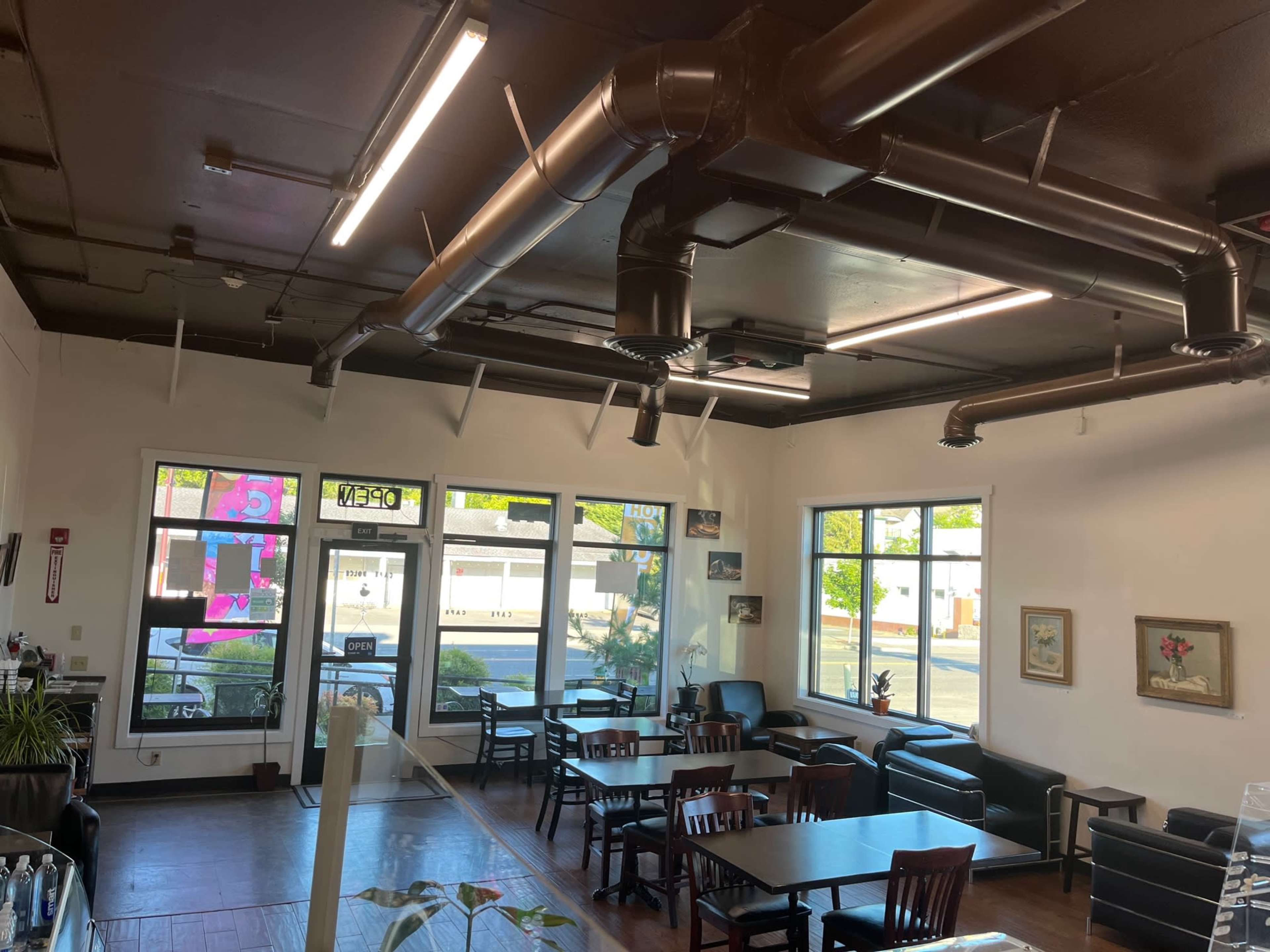 An interior view of a cafe with several tables and chairs, large windows letting in natural light, and exposed ductwork on the ceiling.
