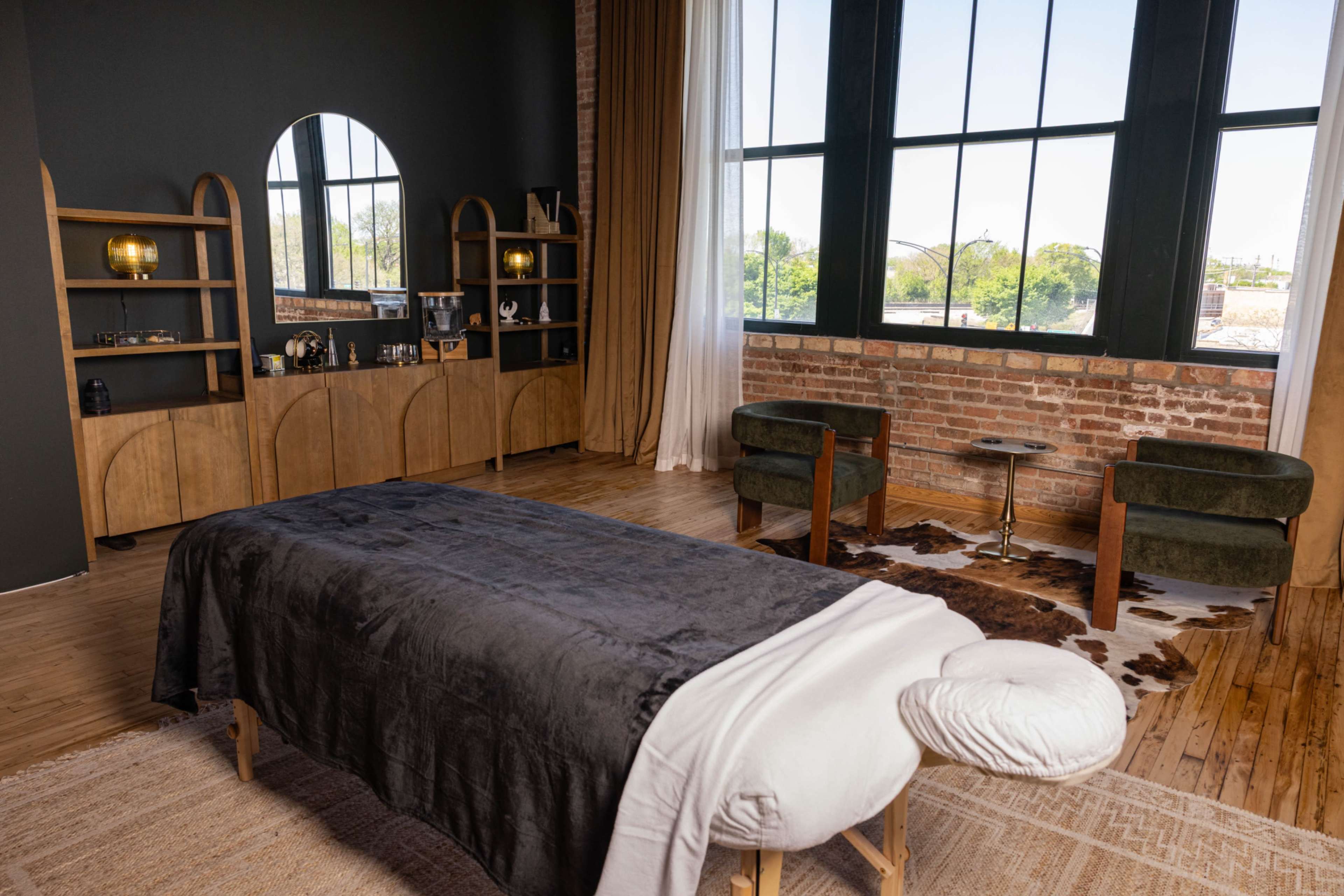 The image shows a minimalist spa room featuring a massage table, two green chairs, and large windows with a view of greenery outside.