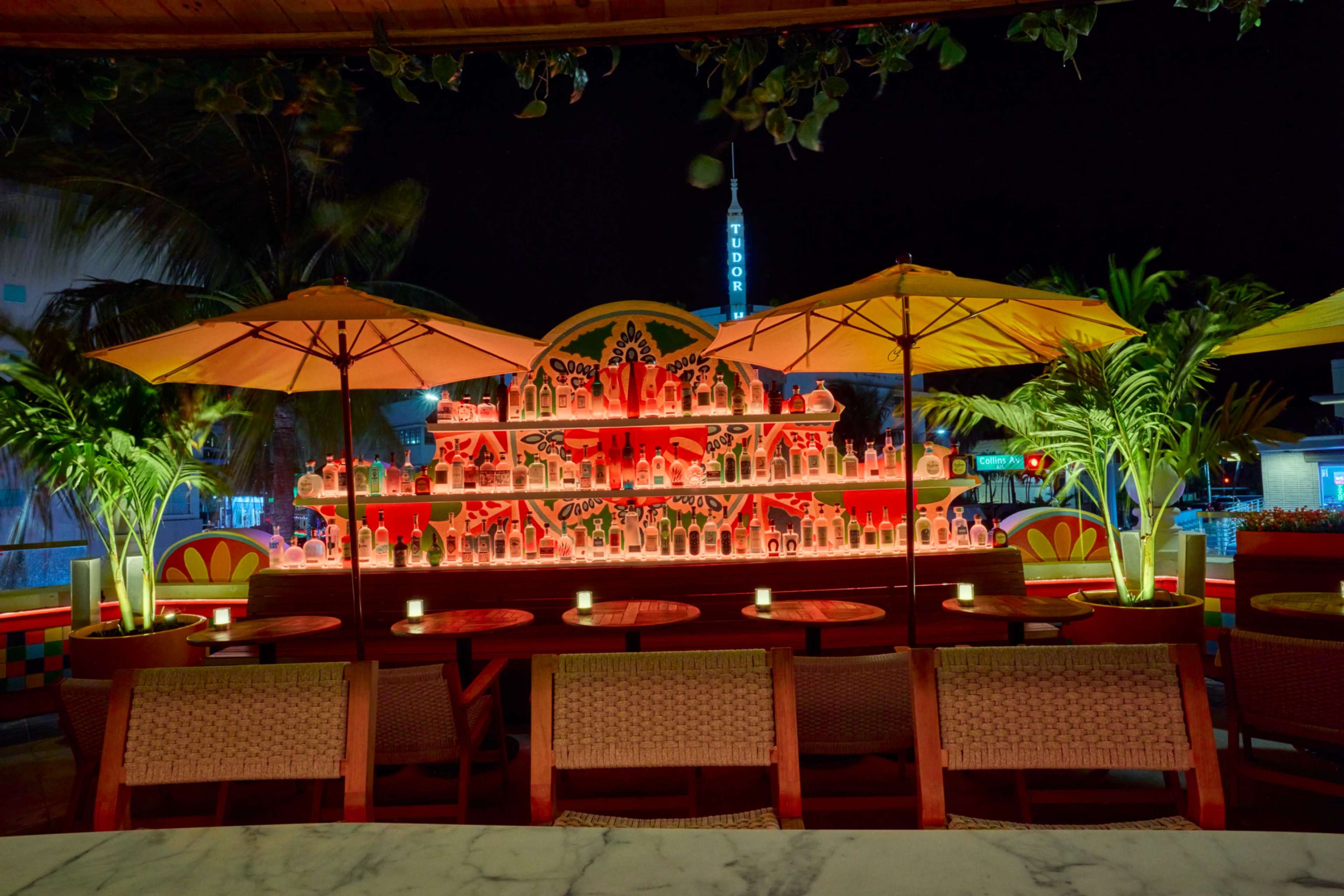 The image shows a brightly lit bar display featuring various bottles, with decorative umbrellas and tropical plants in a nighttime setting.