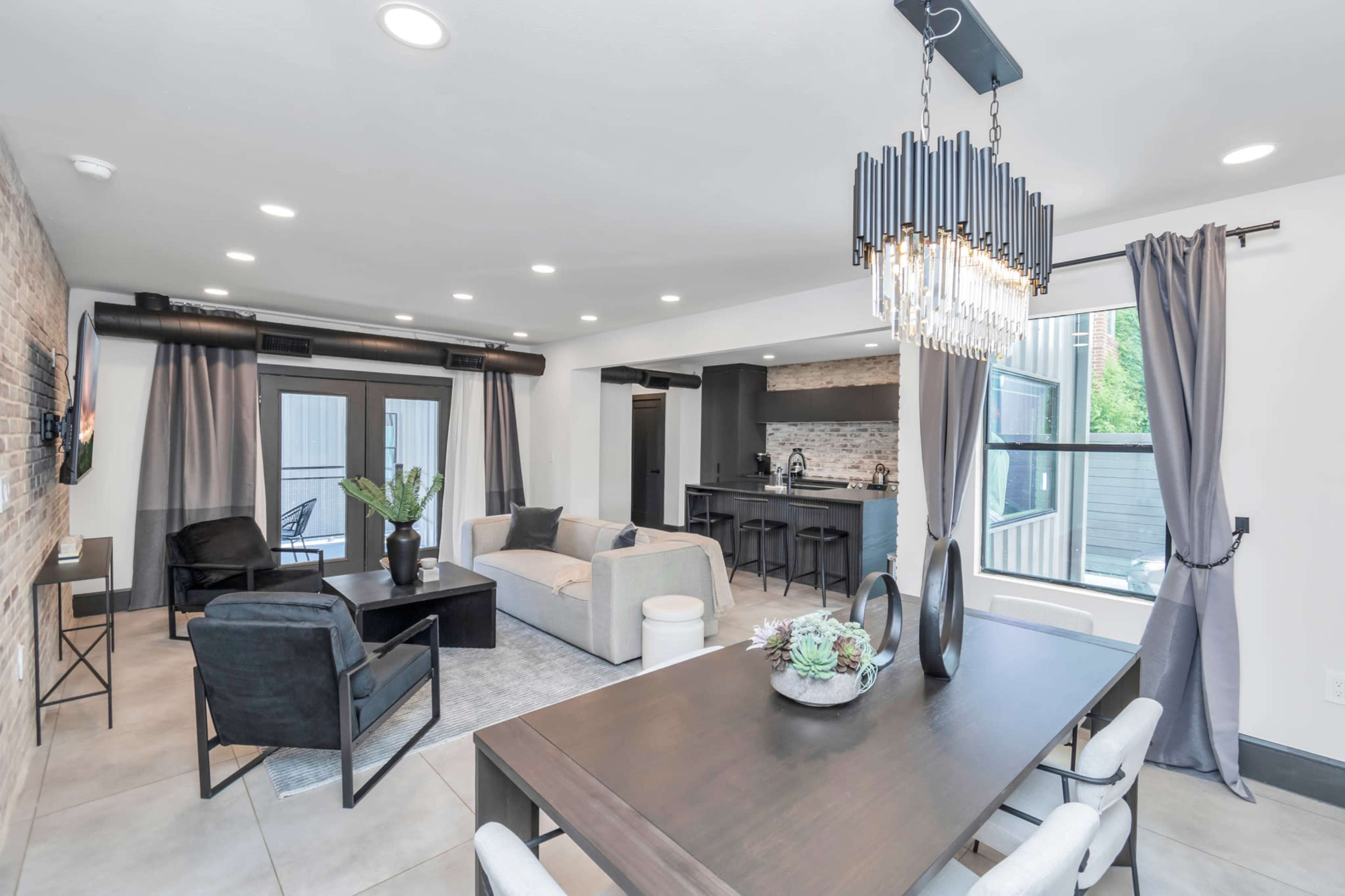 The image features a modern living and dining area with a seating arrangement, a dining table, and large windows letting in natural light.