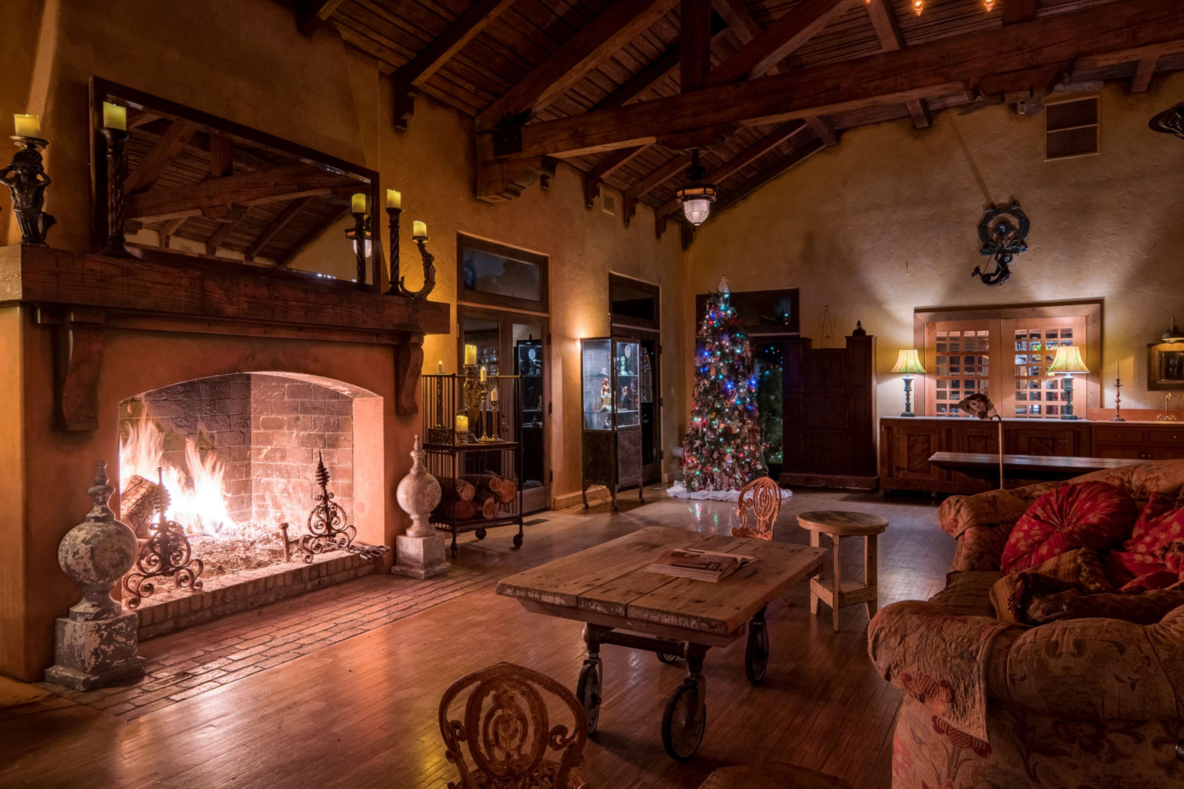 A warmly lit living room features a stone fireplace with a fire burning, a decorated Christmas tree, and rustic wooden furniture.