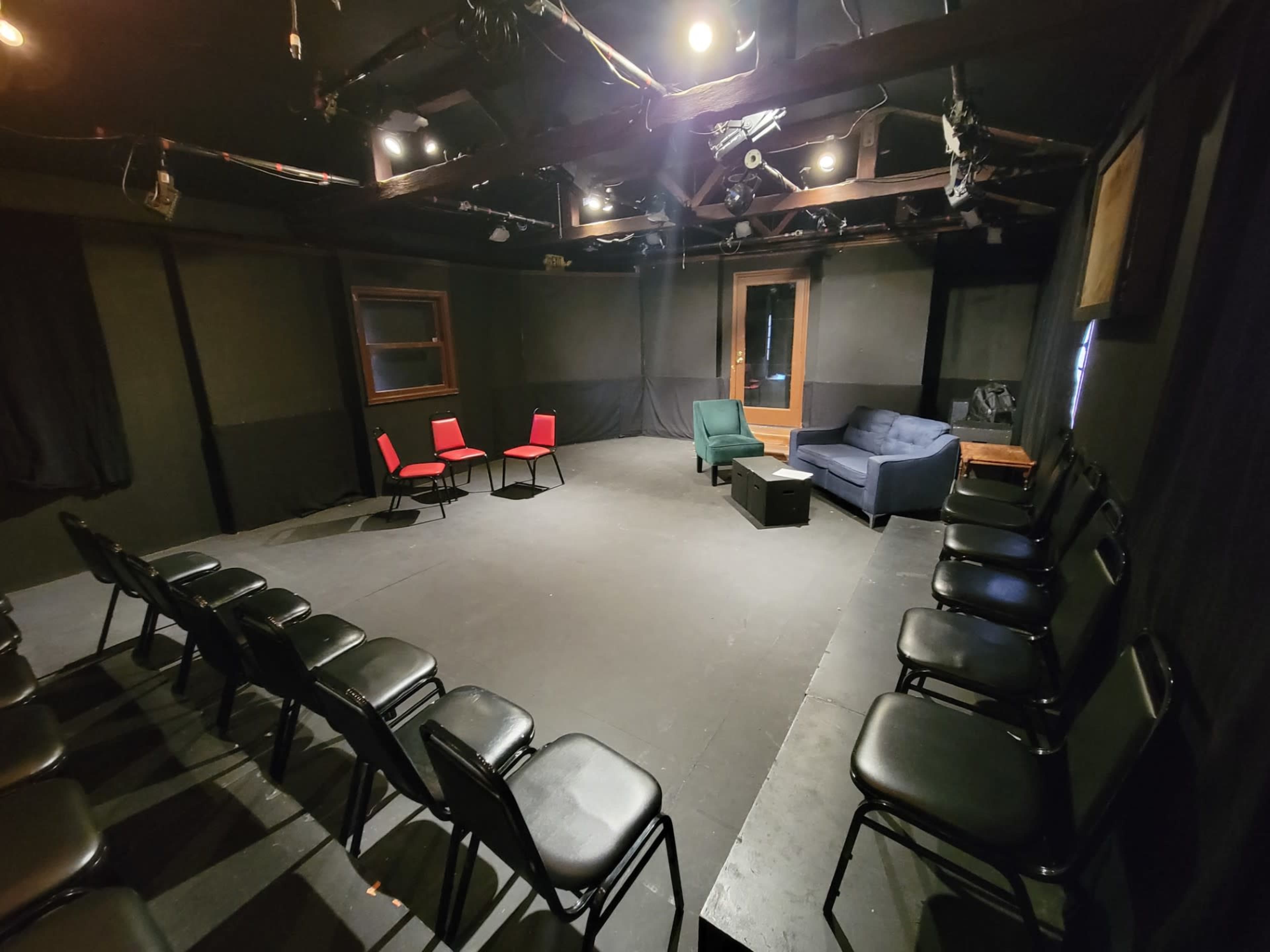 The image shows a small, dimly lit theater space with a circular arrangement of chairs and a few pieces of furniture, including a green chair and a blue sofa.