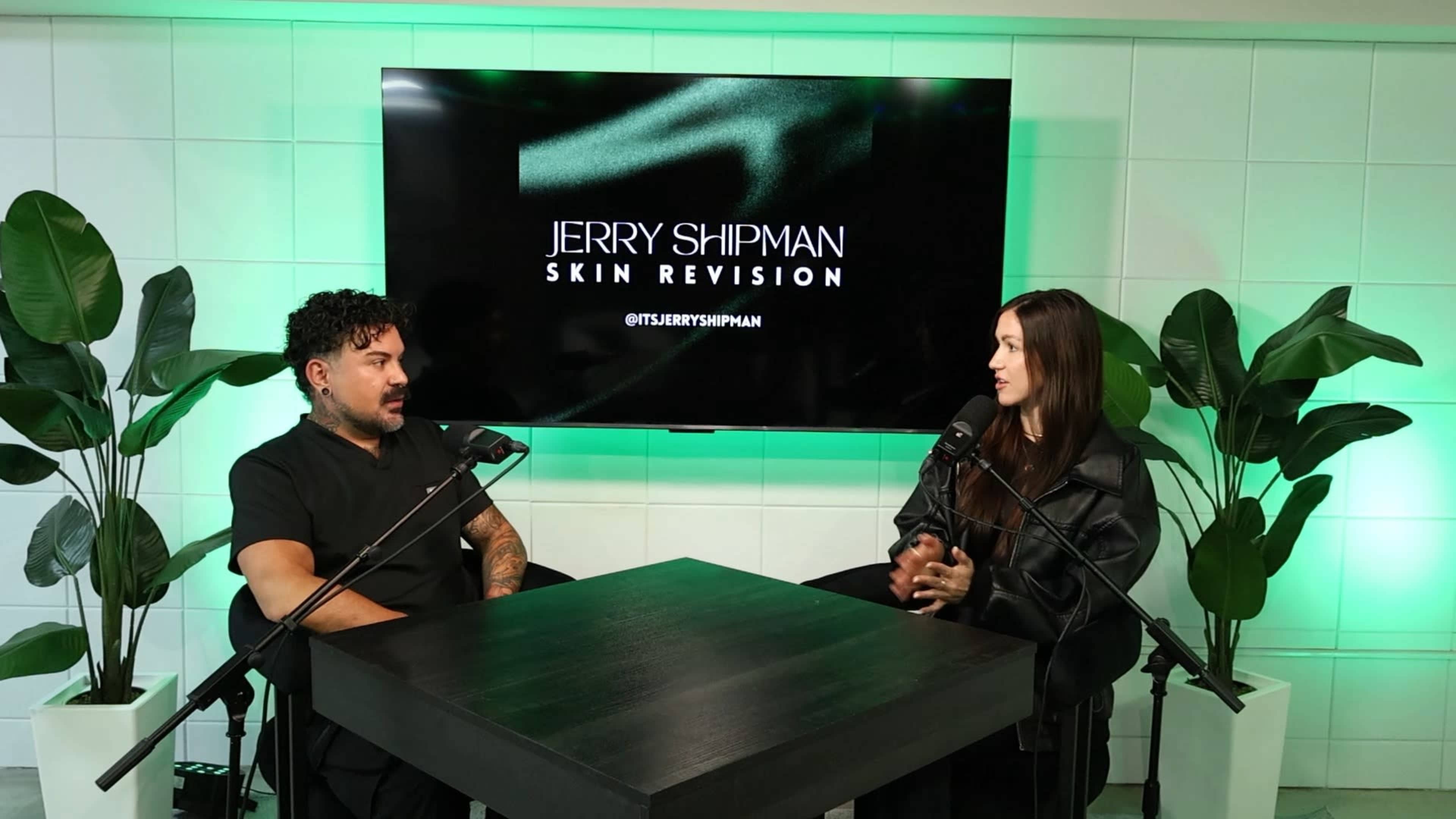 A man and a woman are seated at a table with microphones in front of a large screen displaying "JERRY SHIPMAN SKIN REVISION."
