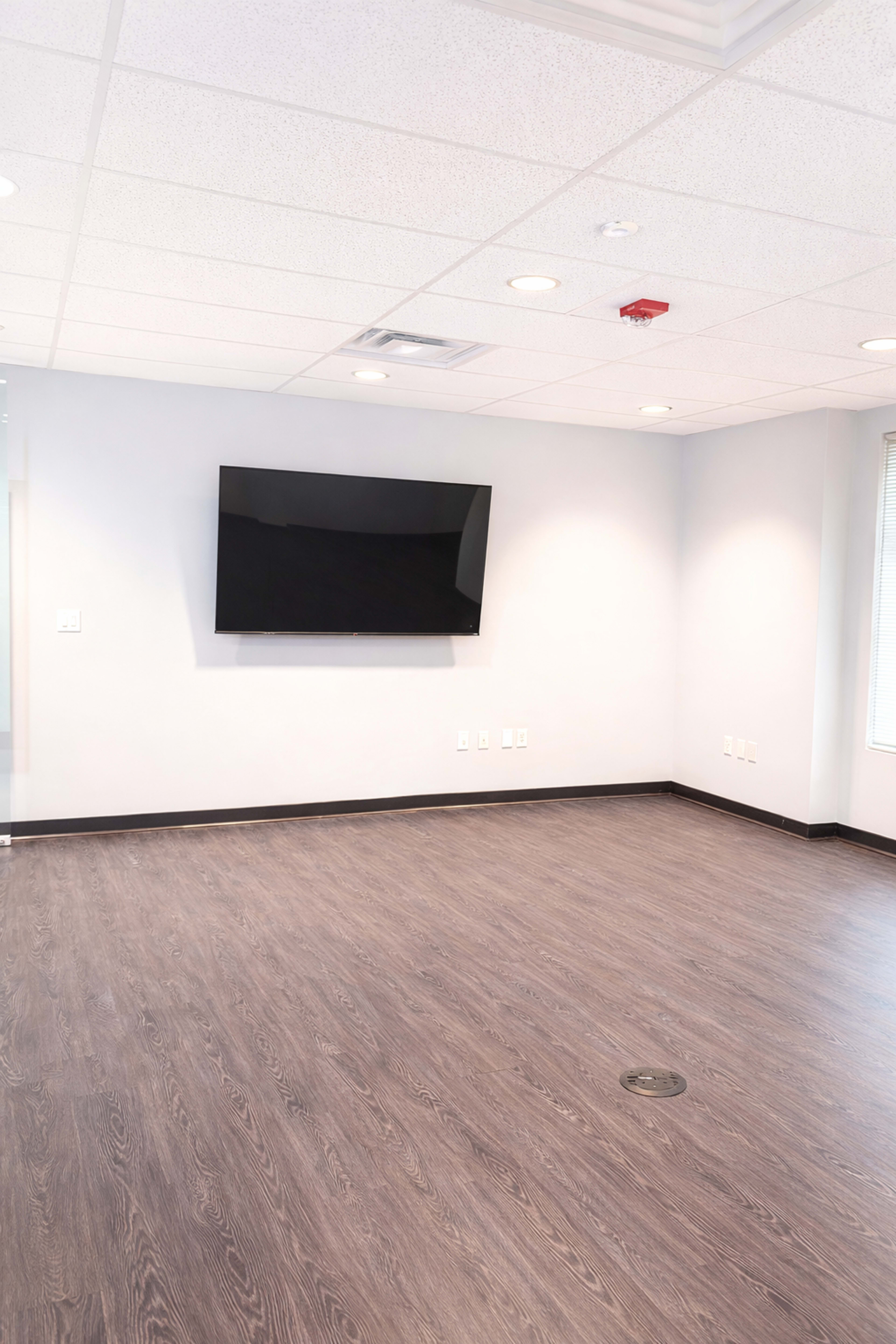 The image shows an empty office space with a television mounted on the wall and a wooden floor.