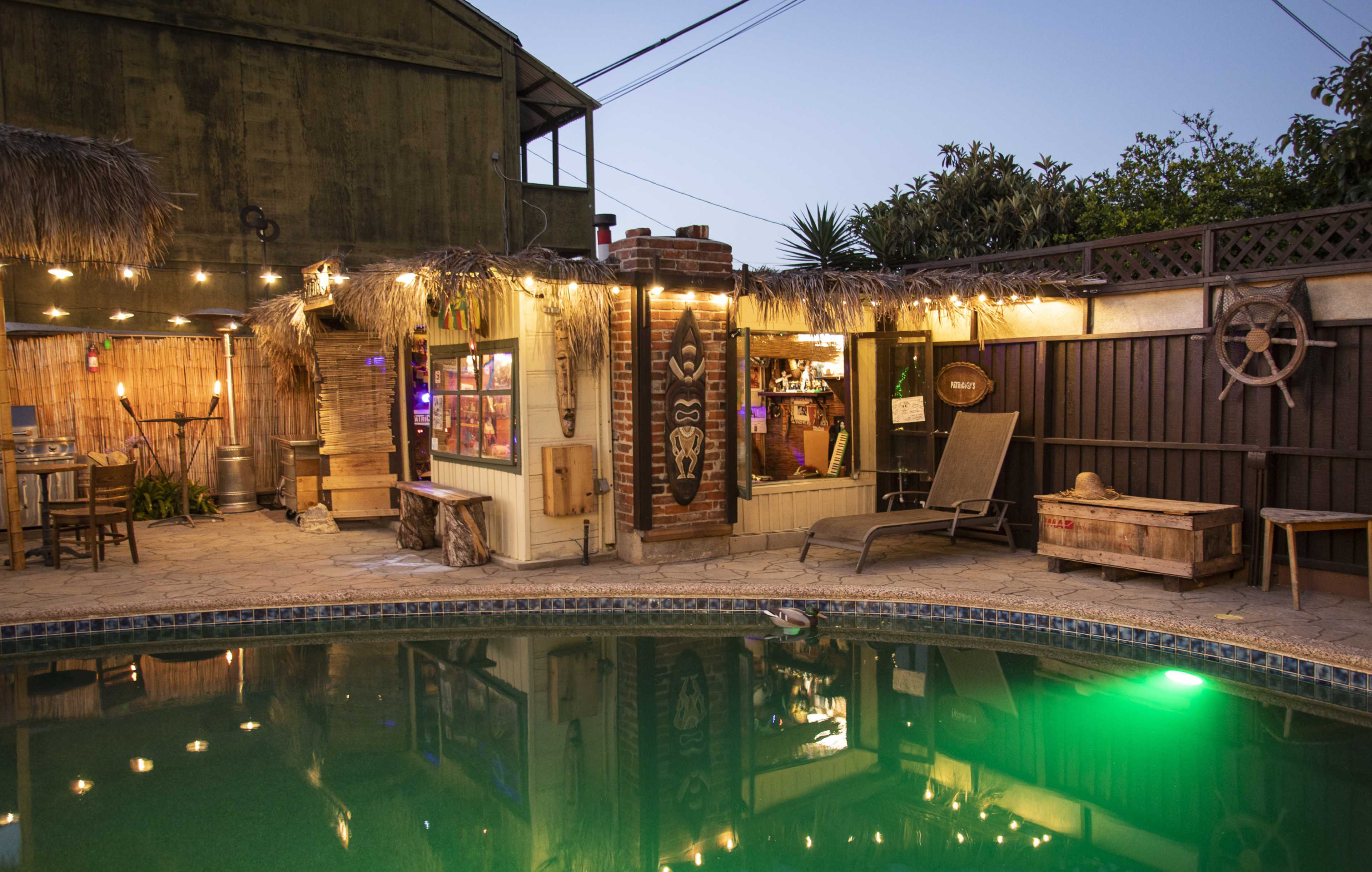 A backyard scene featuring a pool surrounded by a tiki-style bar, string lights, and a seating area within a tropical setting.