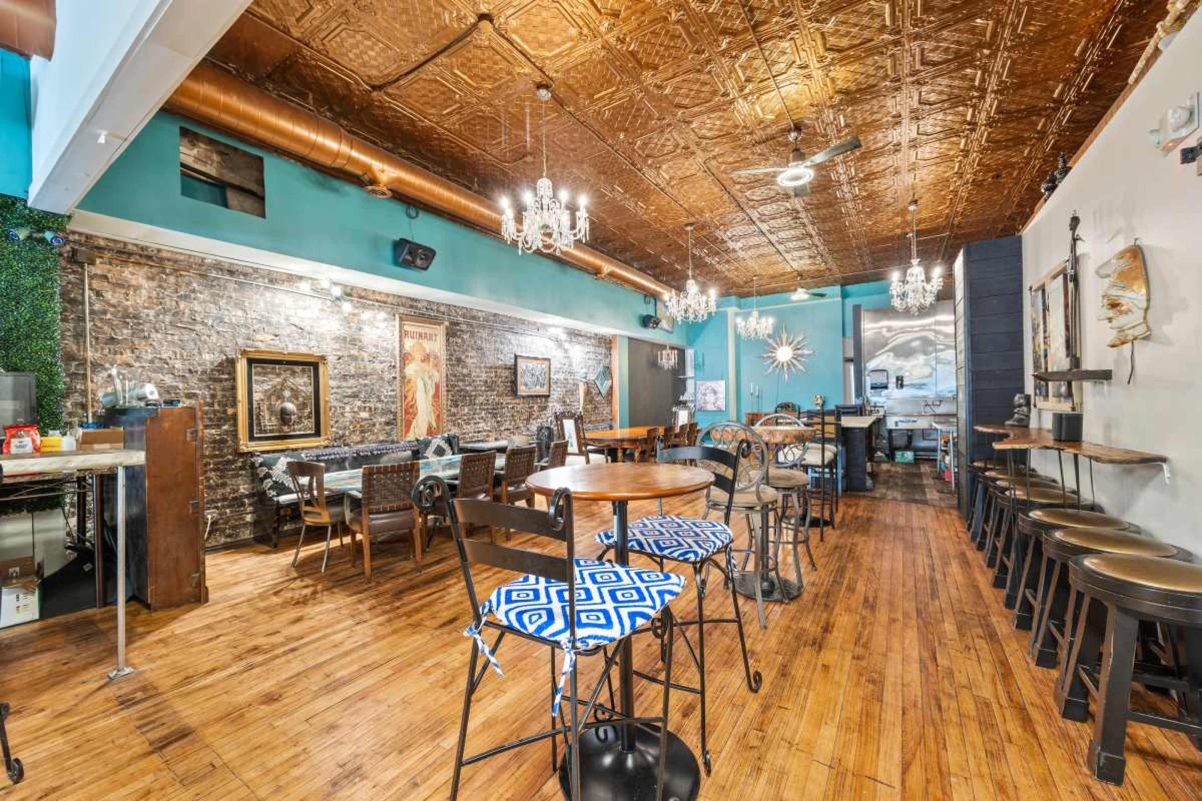 The image shows a dining area with wooden floors, exposed brick walls, and a decorative ceiling, featuring tables and chairs arranged throughout the space.