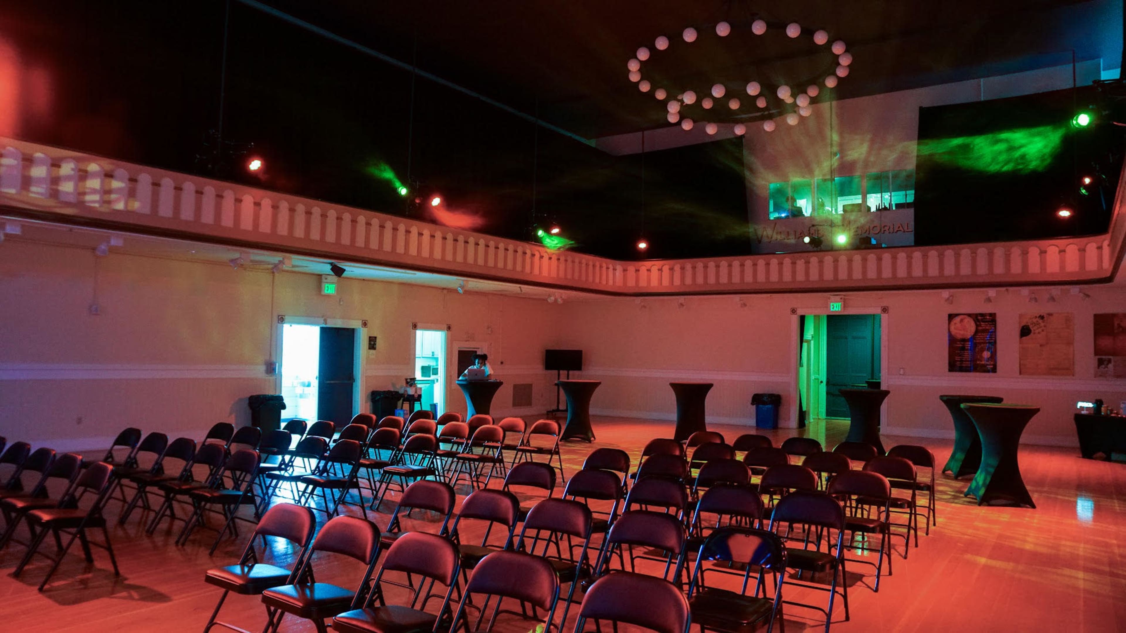 The image shows a spacious event venue featuring rows of empty chairs, with a circular chandelier and colorful lights casting beams across the room.