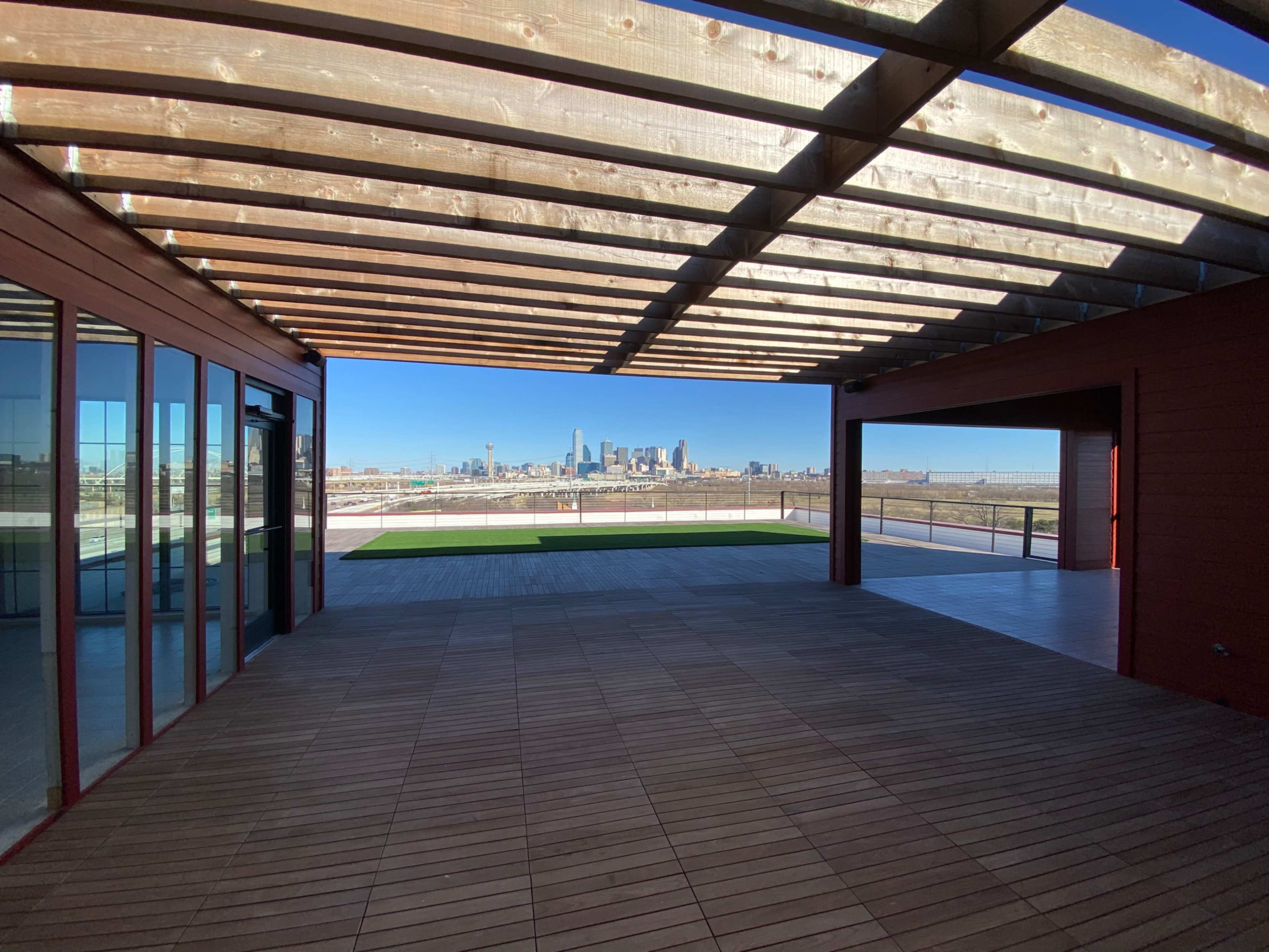 The image shows an open wooden deck with a view of a city skyline through a pergola structure.