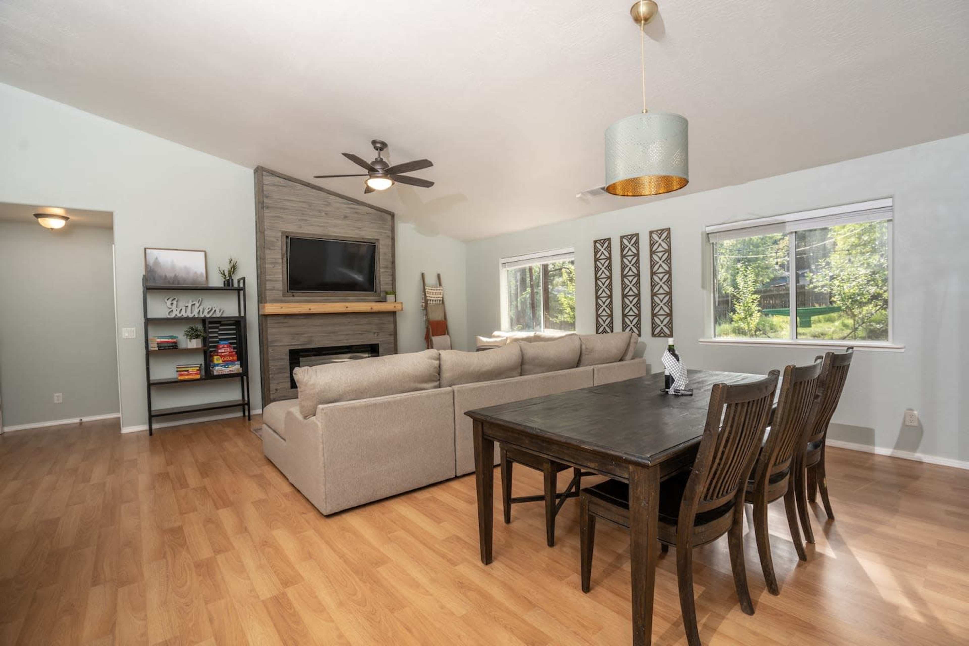 The image shows a spacious living room with a beige sofa, a wooden dining table, and large windows allowing natural light to enter the space.