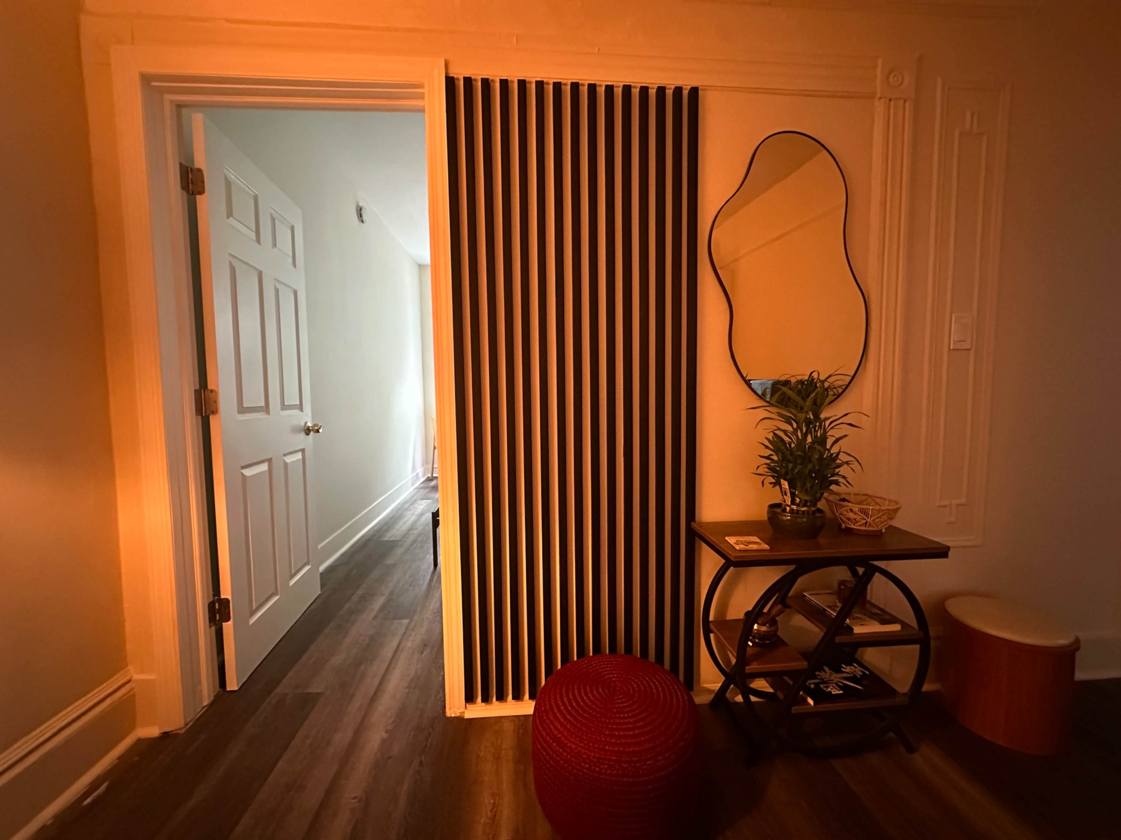 A narrow hallway features a striped wall, a mirror, a potted plant, and a small table with decor items.
