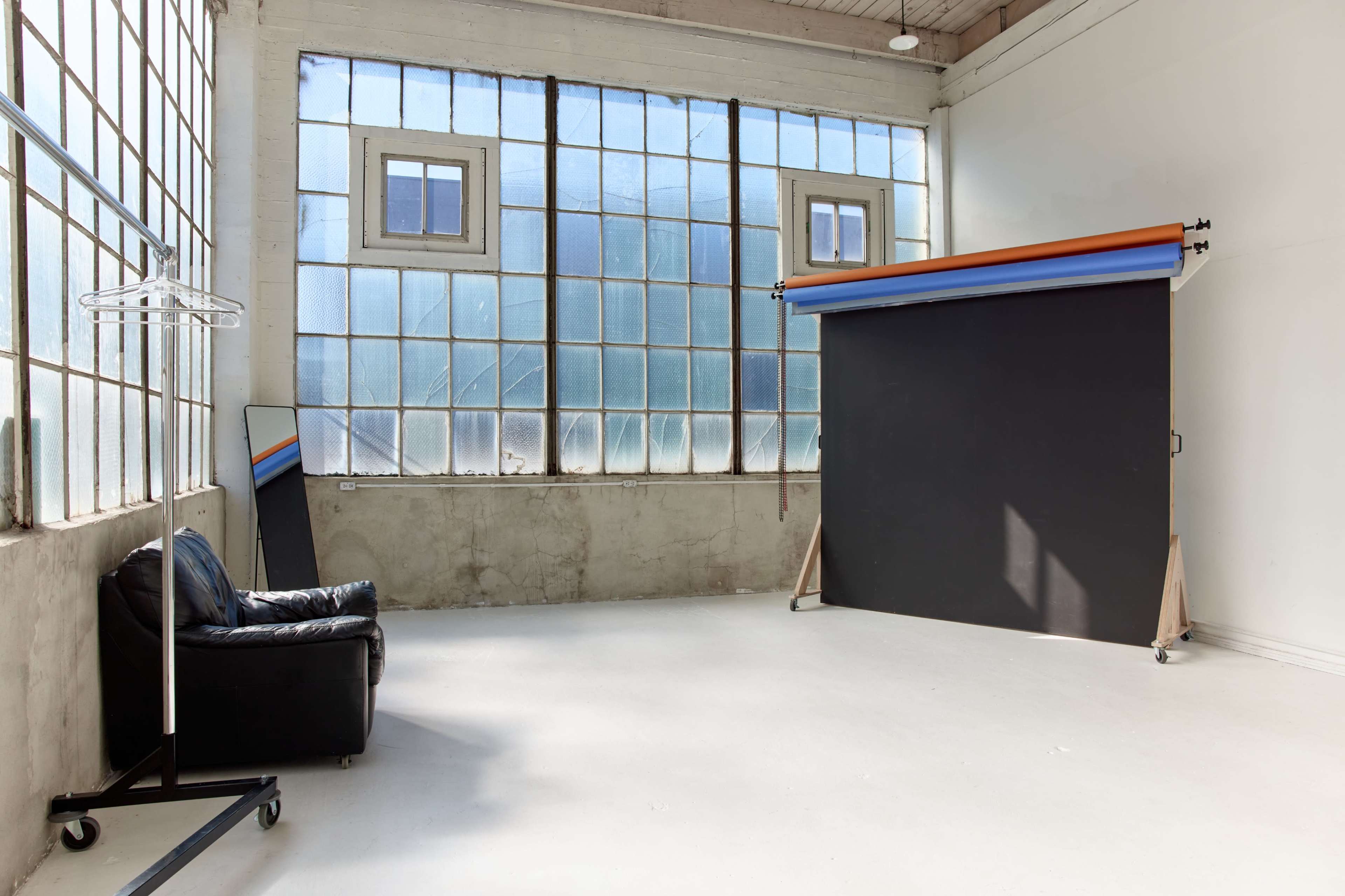 The image shows a spacious photography studio featuring large windows, a black backdrop on stands, a mirror, and a black chair.