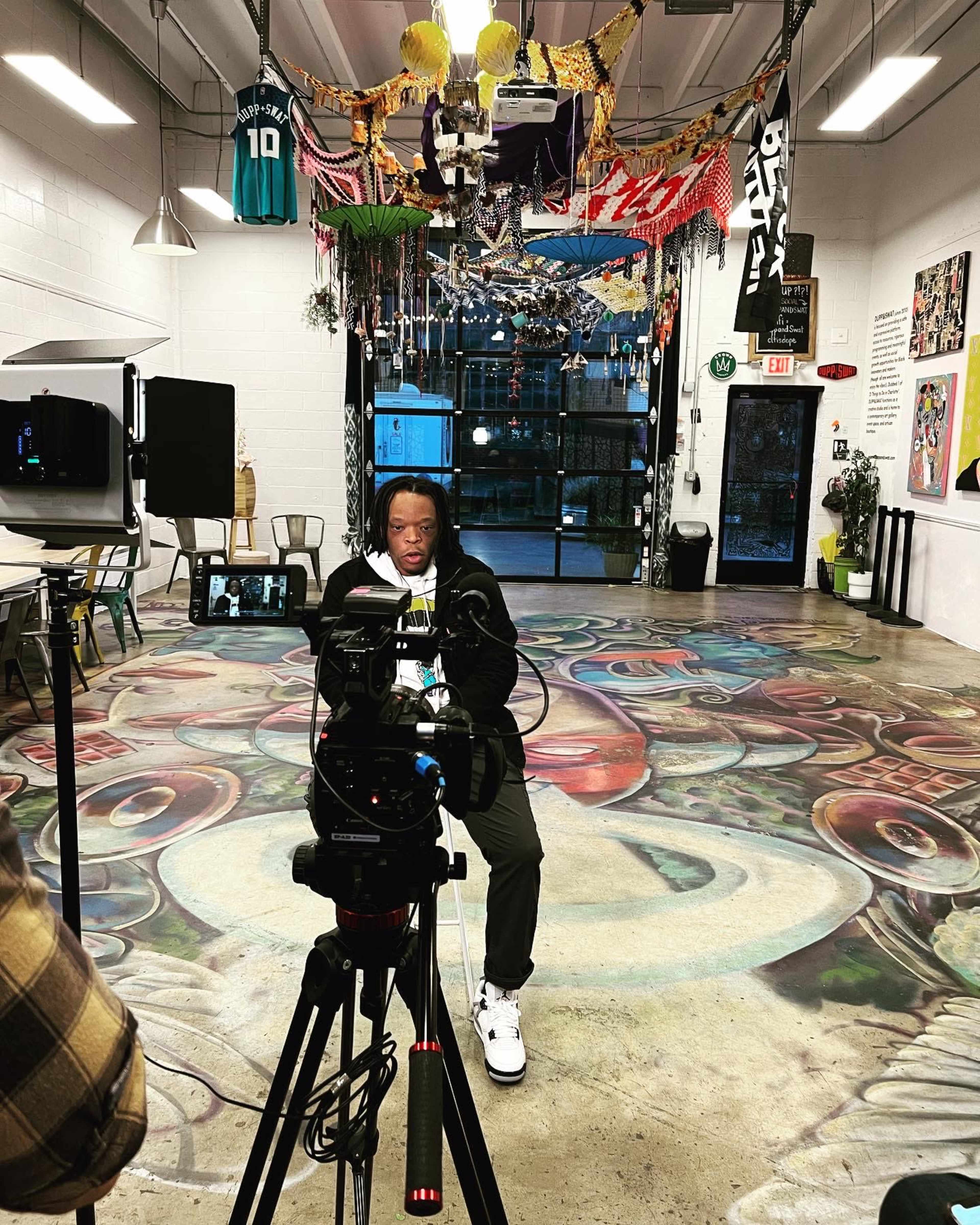 A person sits in front of a camera in a colorful art-filled interior space with a mural on the floor and various decorations hanging from the ceiling.