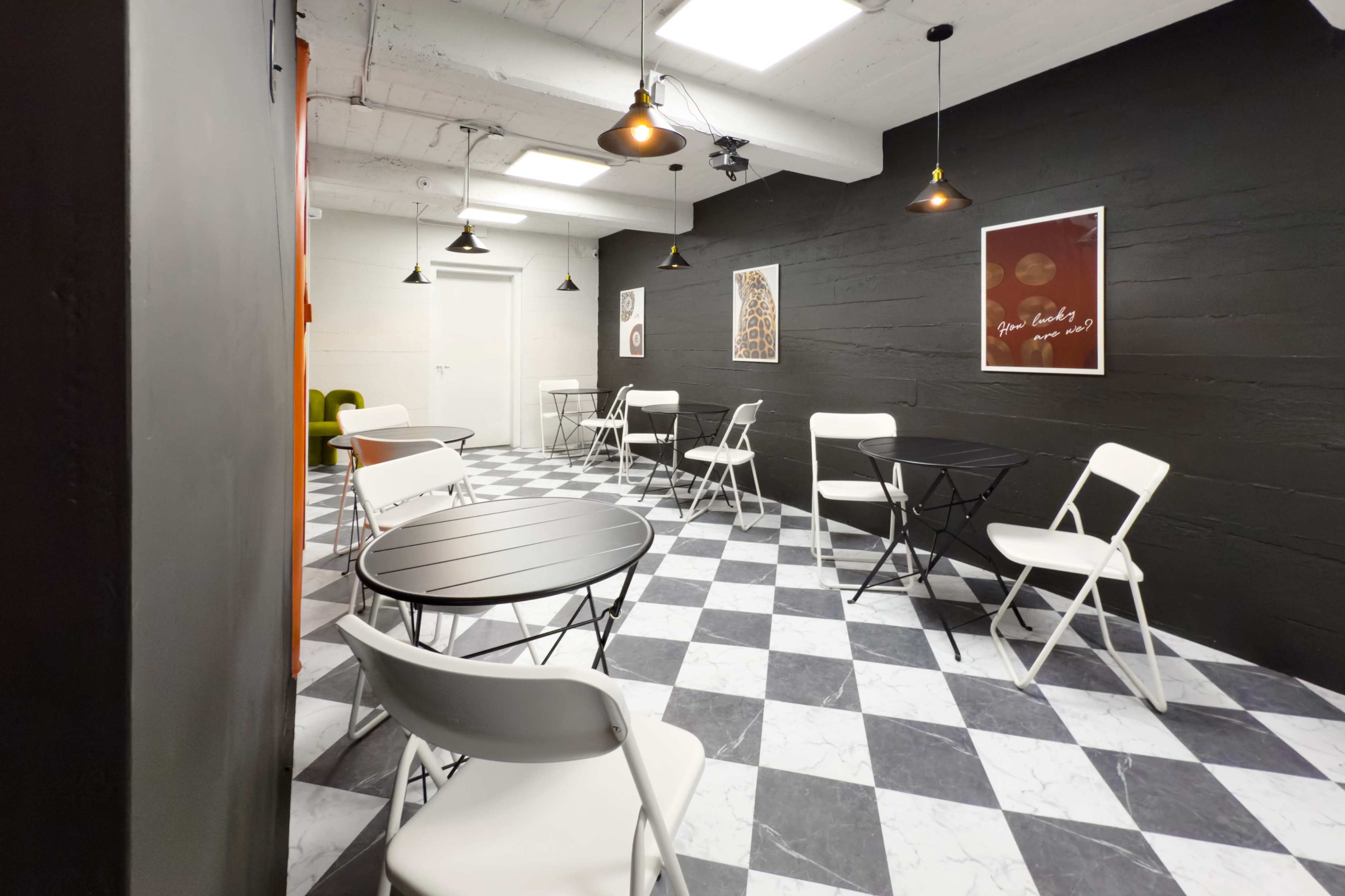 The image shows a modern cafe interior with black and white checkered flooring, several round tables, and white folding chairs against a dark wall.