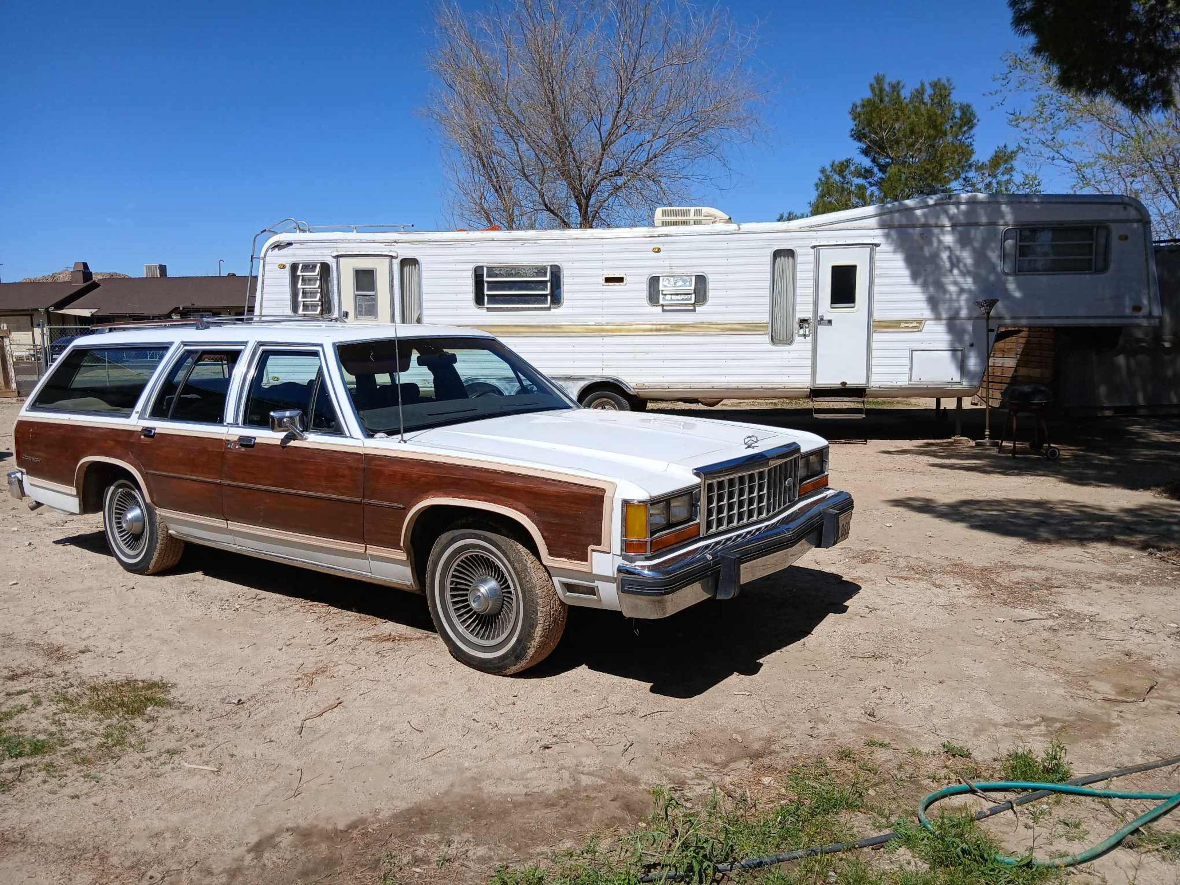 A white and wood-paneled station wagon is parked in front of a vintage travel trailer on a dirt lot.