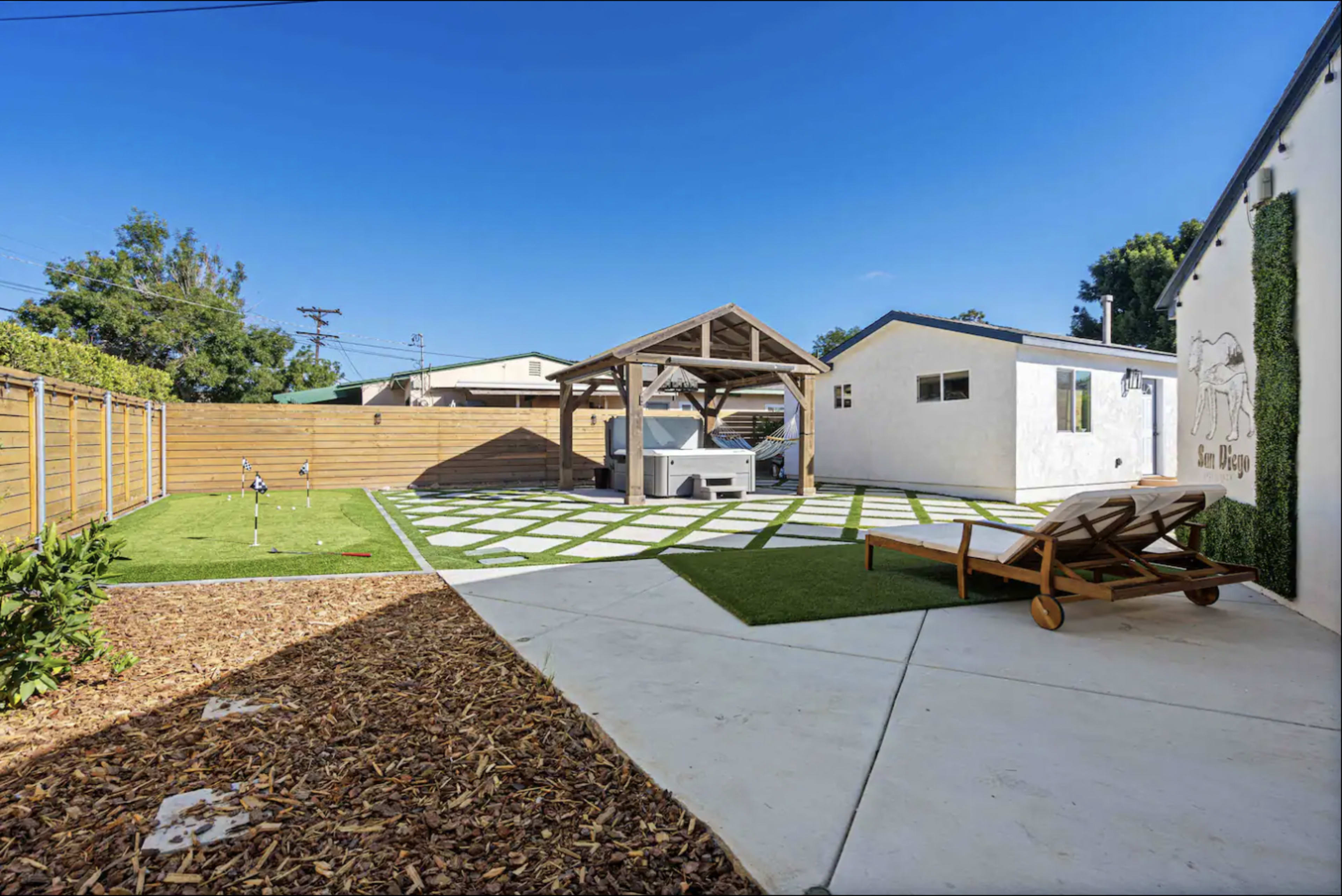 A backyard with a wooden gazebo, a patch of artificial grass, and a concrete patio area with lounge chairs.