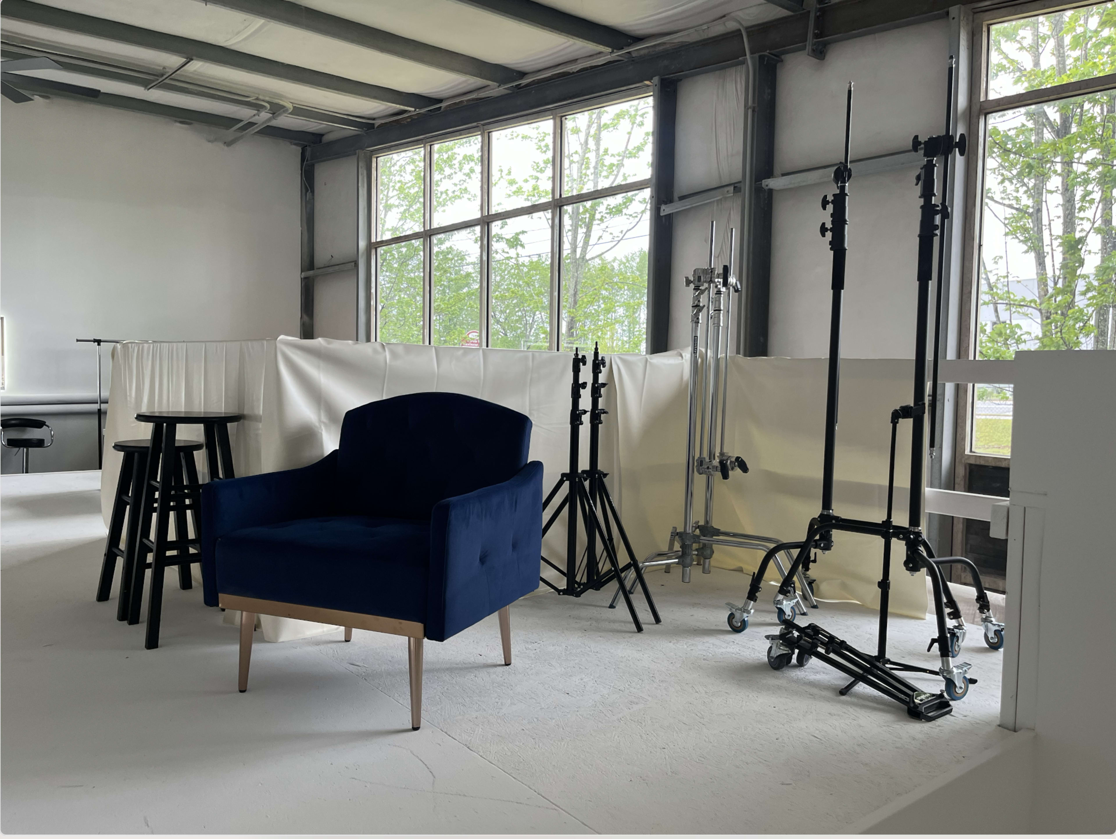 A blue velvet armchair surrounded by studio lighting stands and a tall stool in a spacious room with large windows.