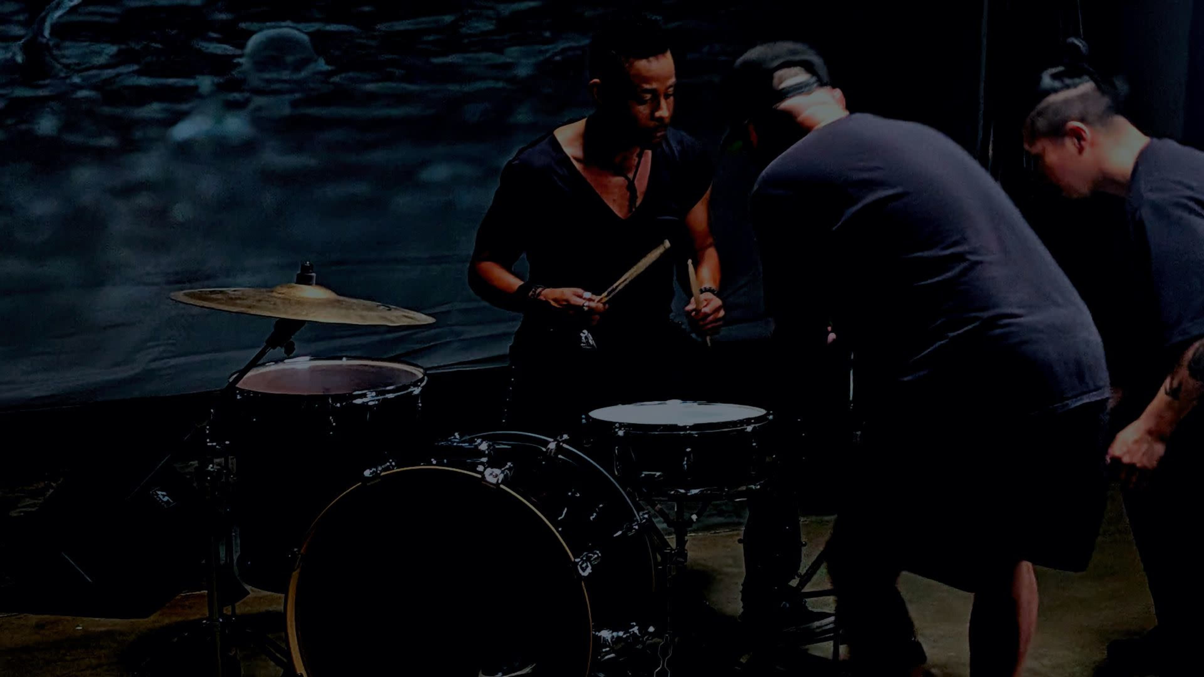 Two musicians are adjusting a drum set in a dimly lit space, with one holding drumsticks and the other focused on the equipment.