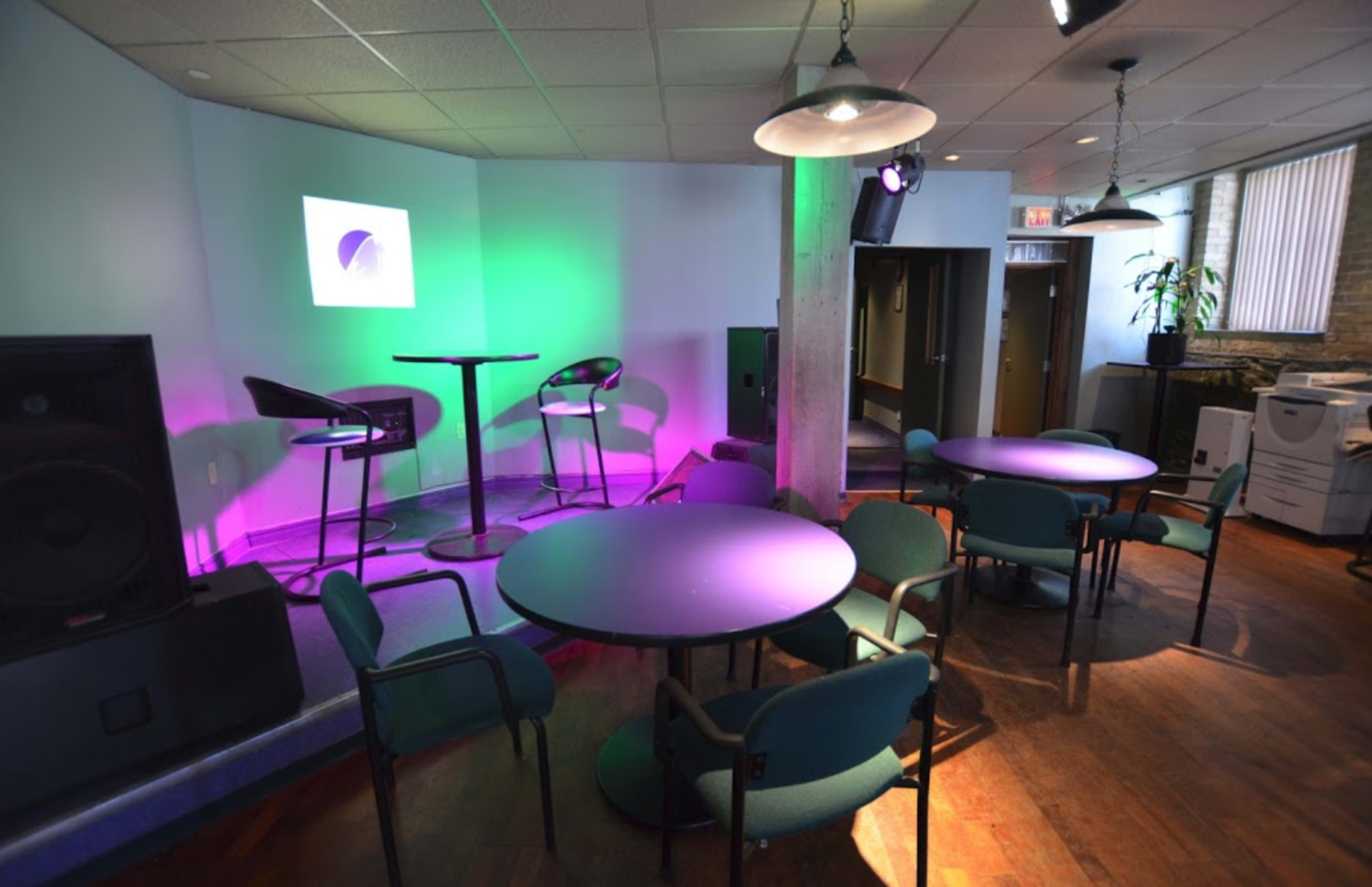 The image shows a small lounge area featuring round tables and chairs, a stage with a colorful light setup, and a projector screen against the wall.