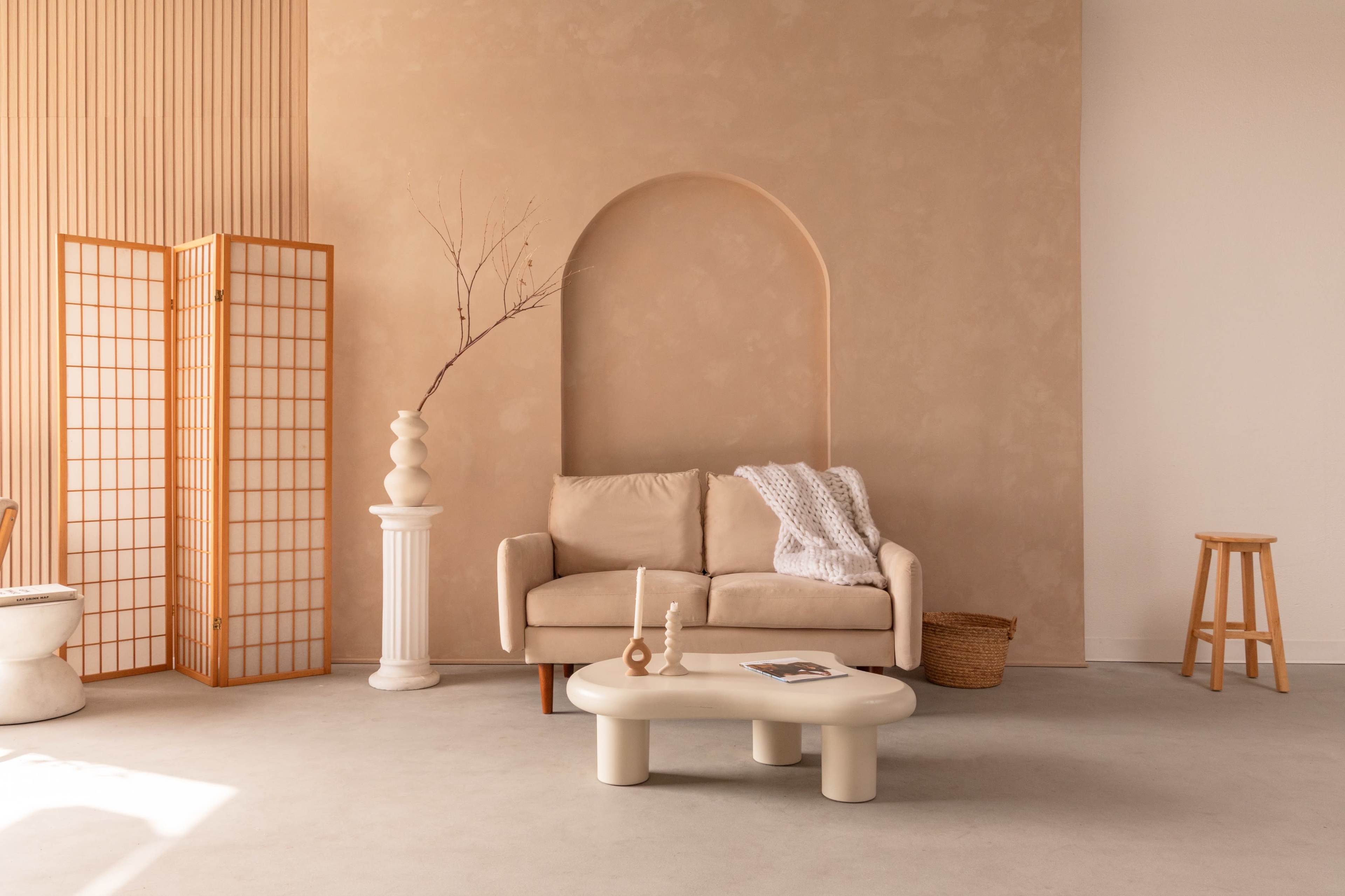 The image features a minimalist living space with a beige sofa, a round coffee table, and a tall vase on a pedestal, set against a softly textured wall and a wooden screen.