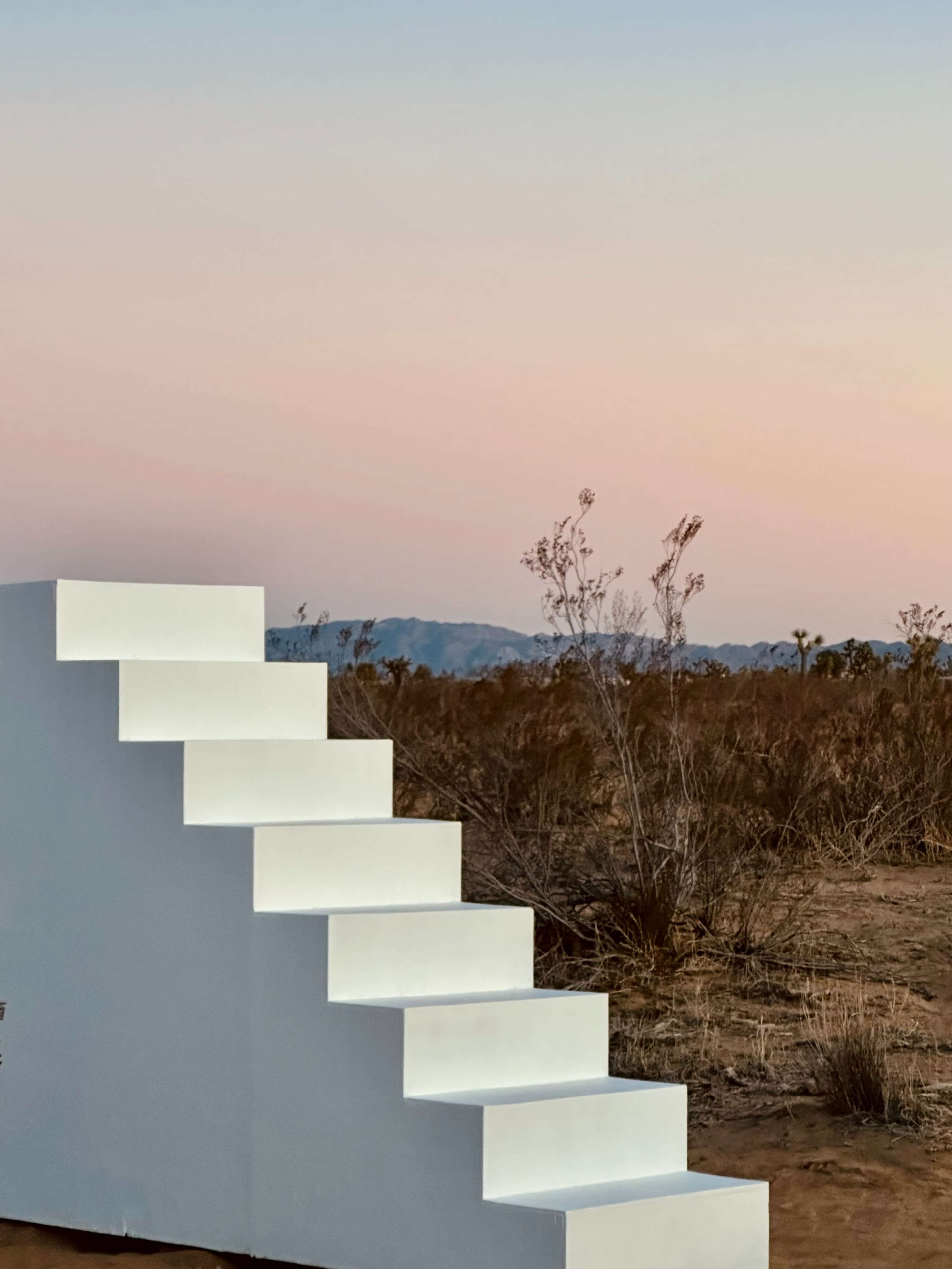 The image features a white staircase set against a desert landscape with soft hues in the sky and distant mountains.
