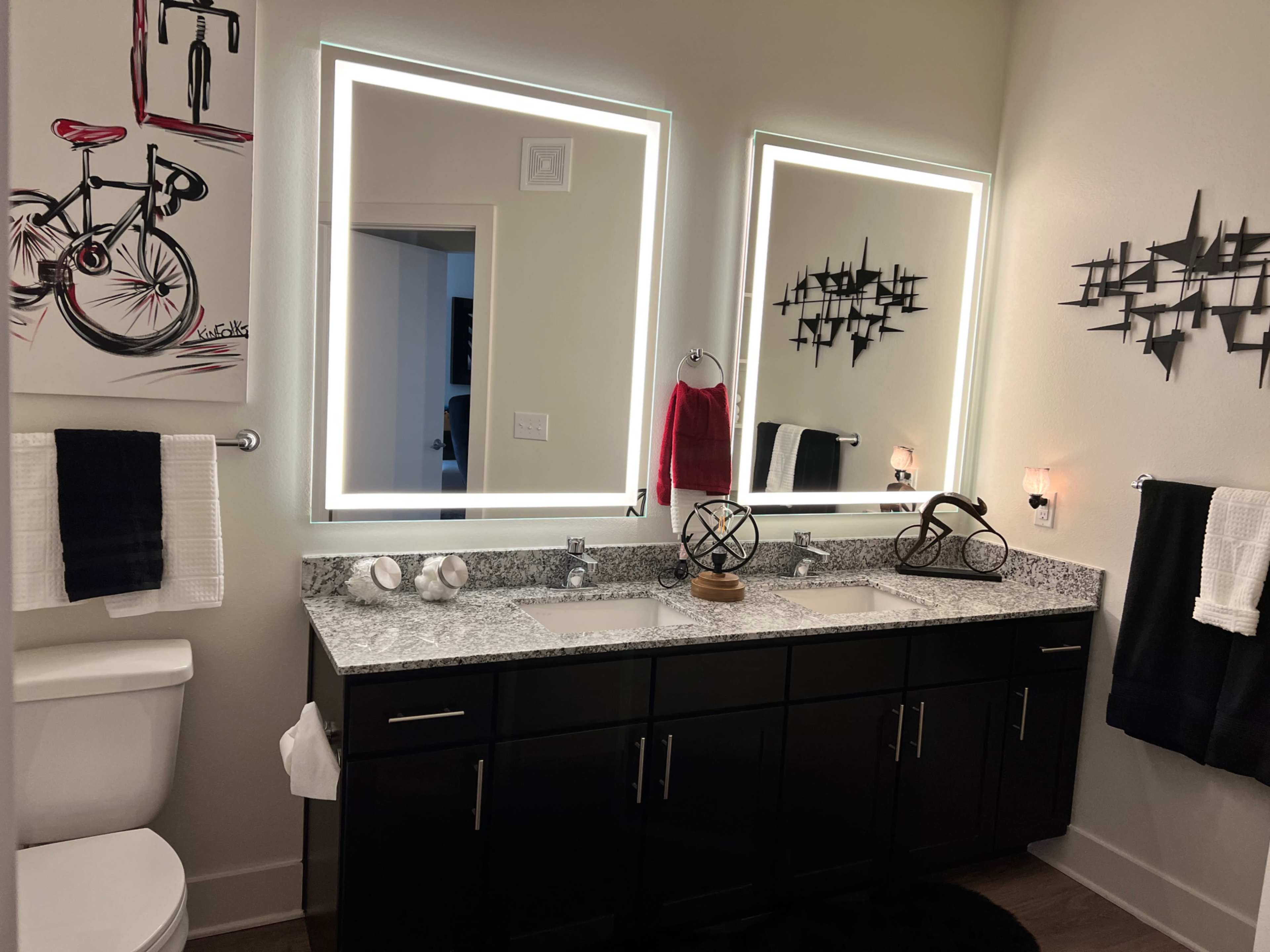 A modern bathroom features dual sinks with a granite countertop, illuminated mirrors, and a decorative bicycle print on the wall.