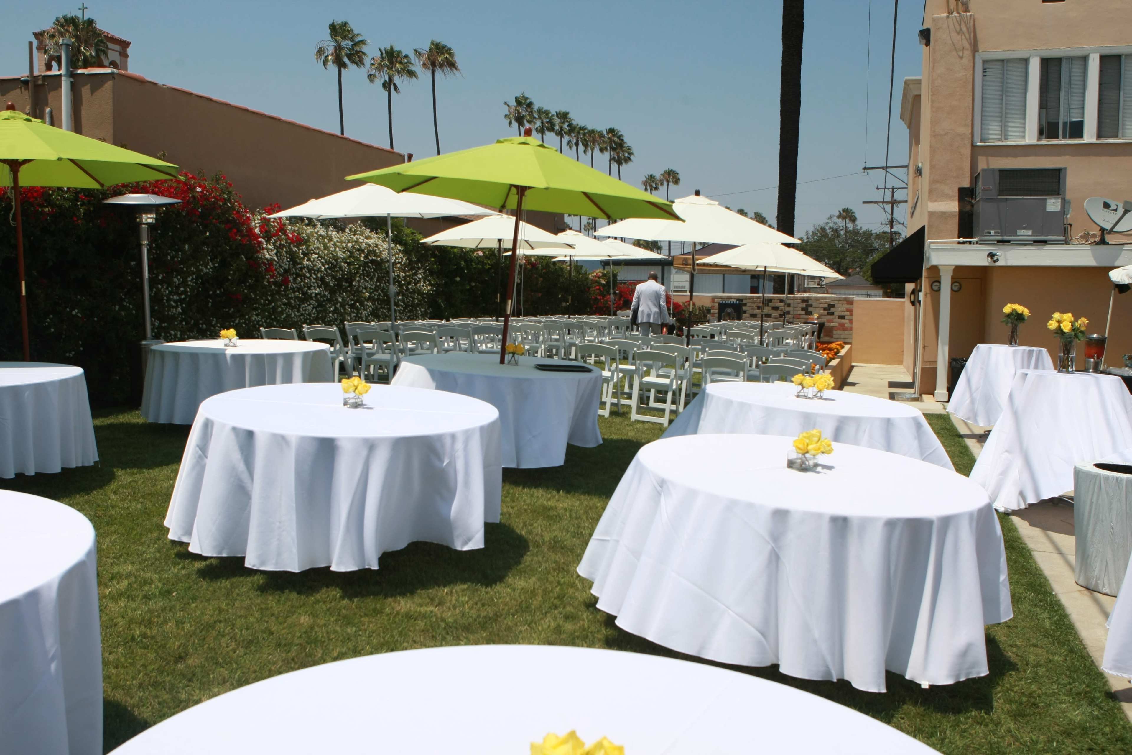 The outdoor setting features several round tables with white tablecloths and yellow flower centerpieces, shaded by green umbrellas, against a backdrop of palm trees.