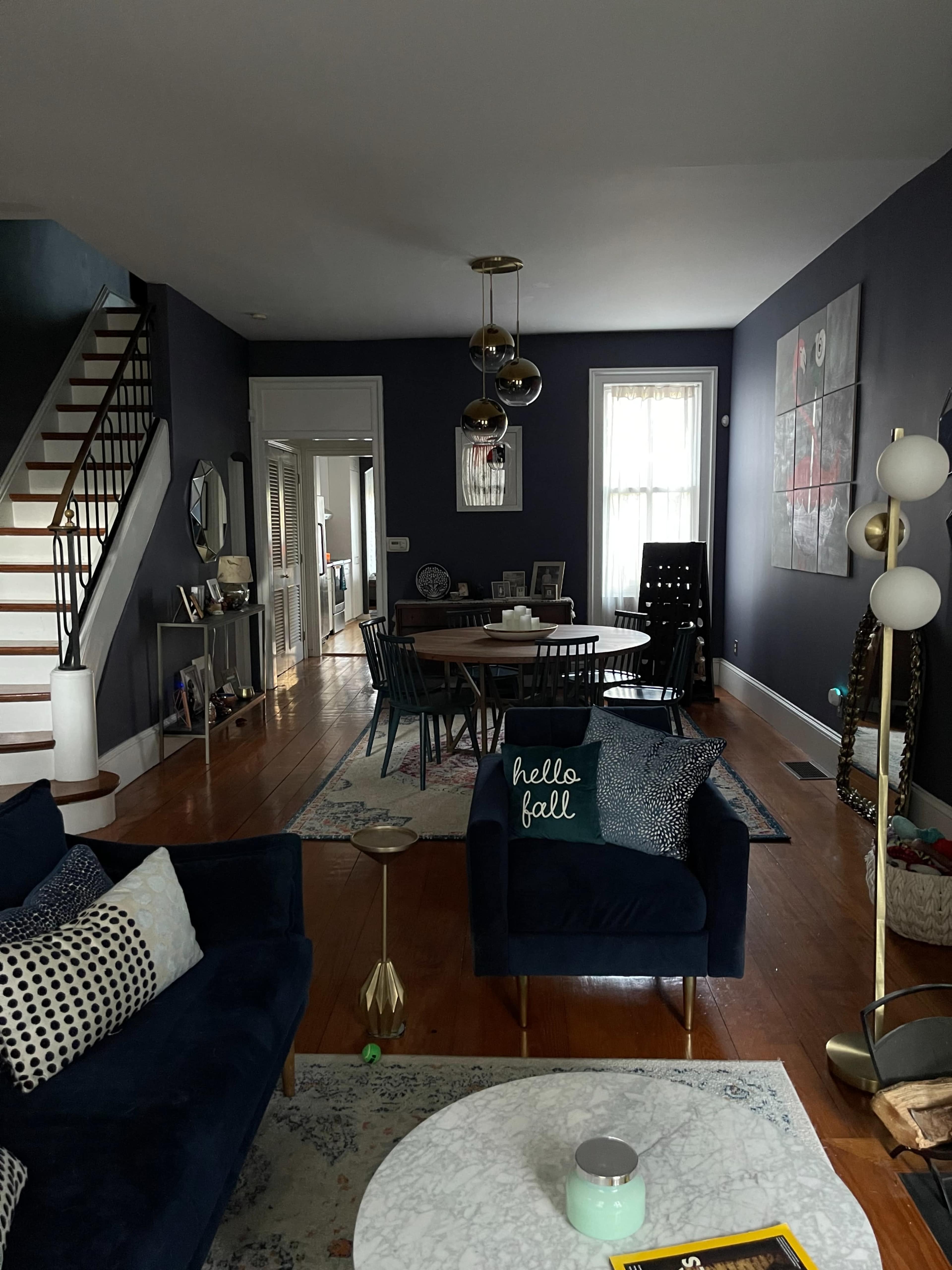 A living room with a dark blue wall, a seating area with a navy couch and patterned cushions, a dining table in the background, and a staircase leading to an upper floor.