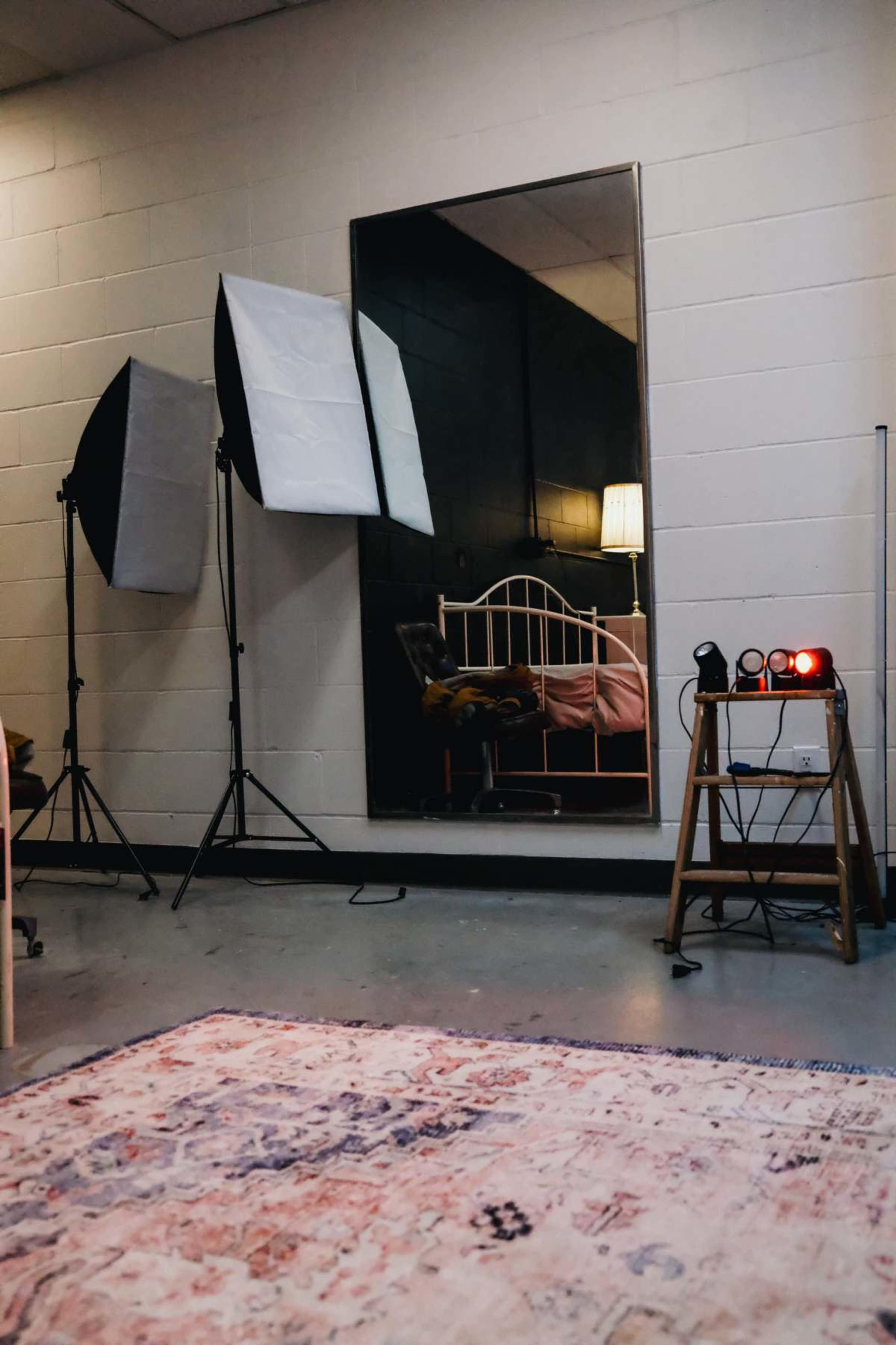 The image shows a room with a large mirror, two lighting setups on stands, a bed with pink bedding, and a ladder with lights positioned next to it.