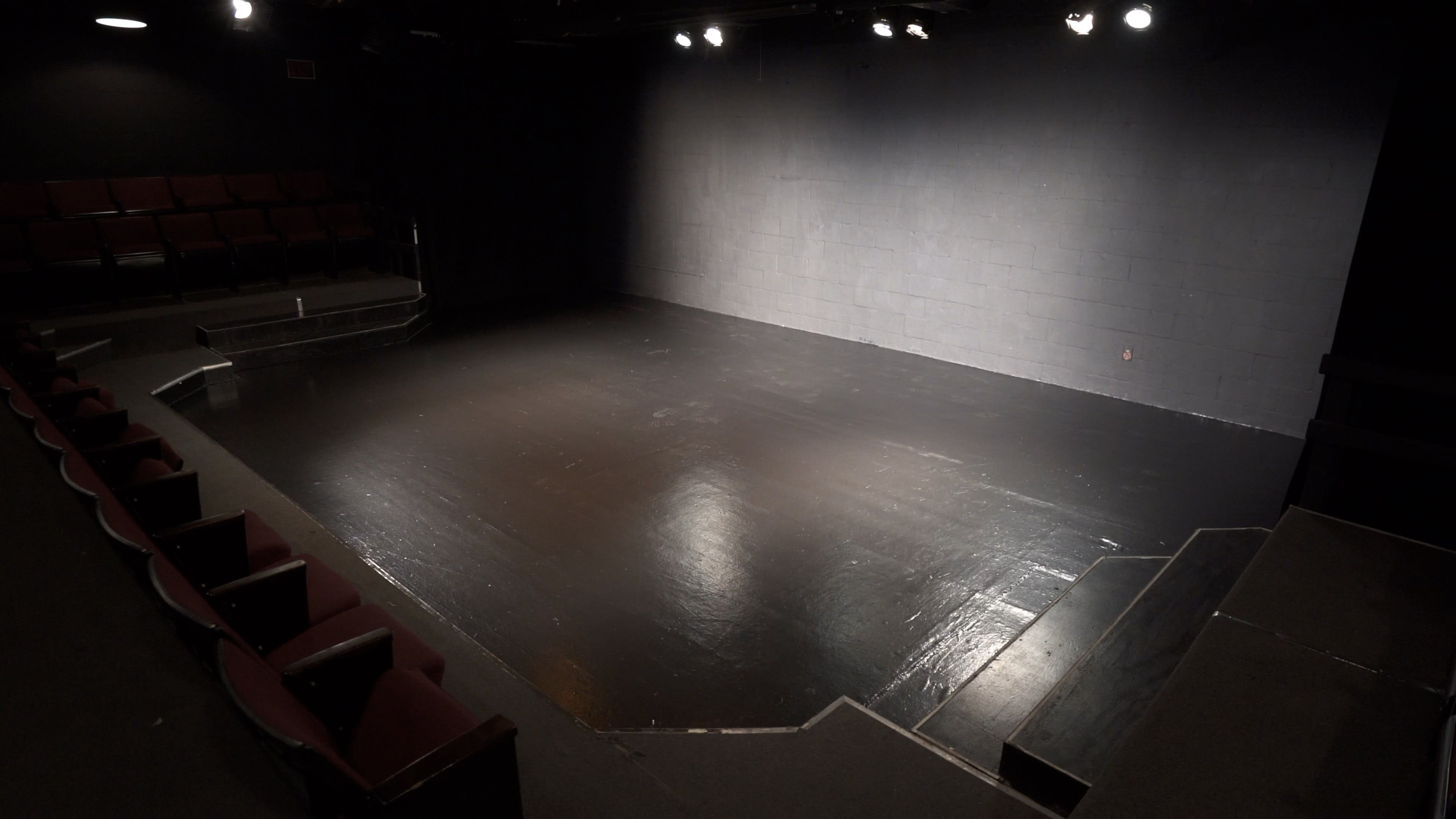 A vacant theater stage with a black floor and a dark backdrop, illuminated by overhead lights, and featuring empty rows of red seating.