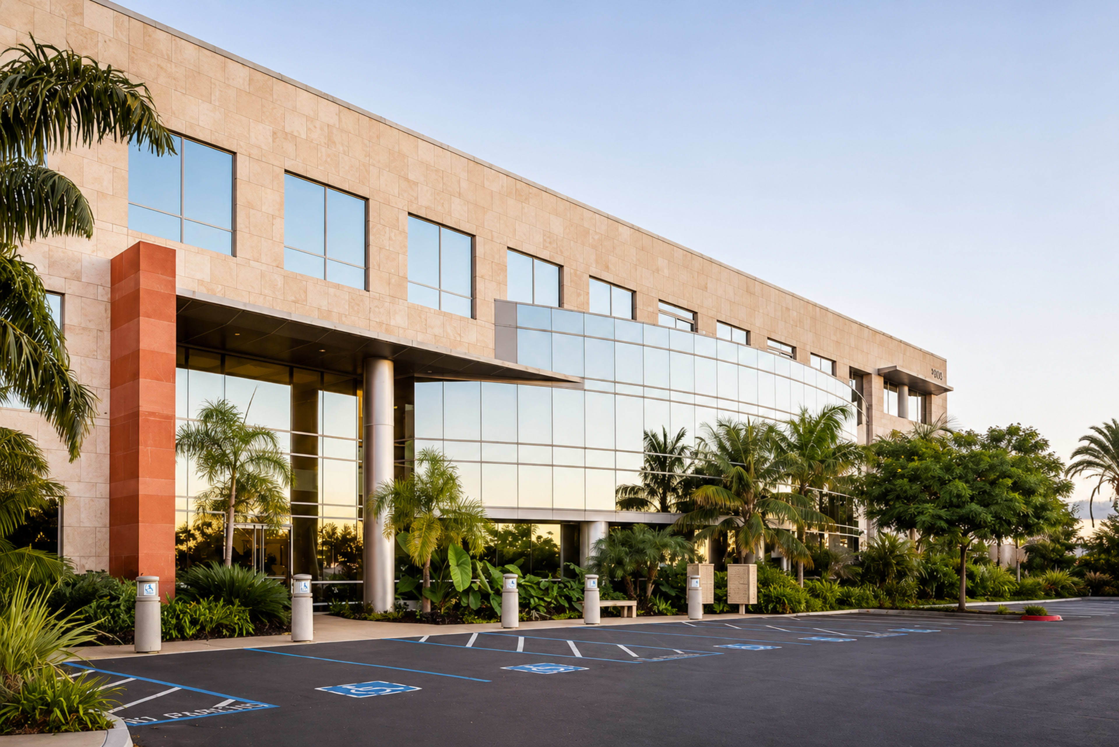The image shows a modern three-story office building with large glass windows, surrounded by tropical landscaping and a parking lot with marked spaces.