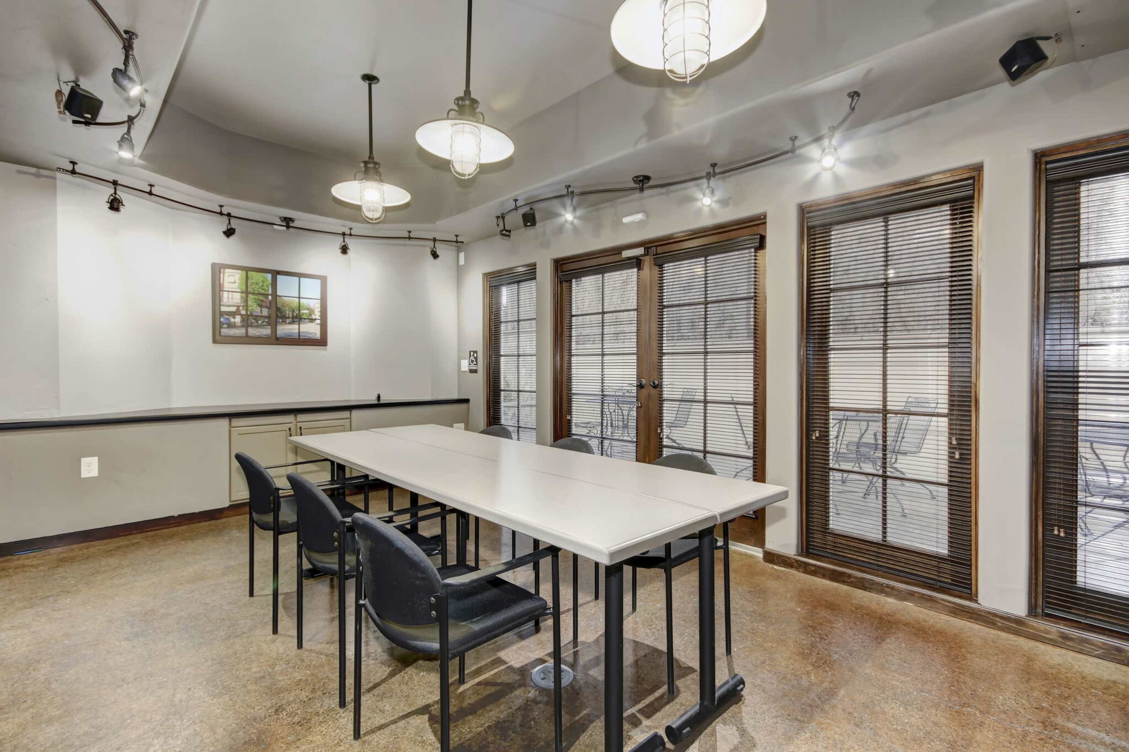 A meeting room with a long table, black chairs, and large windows with blinds, illuminated by pendant lights.
