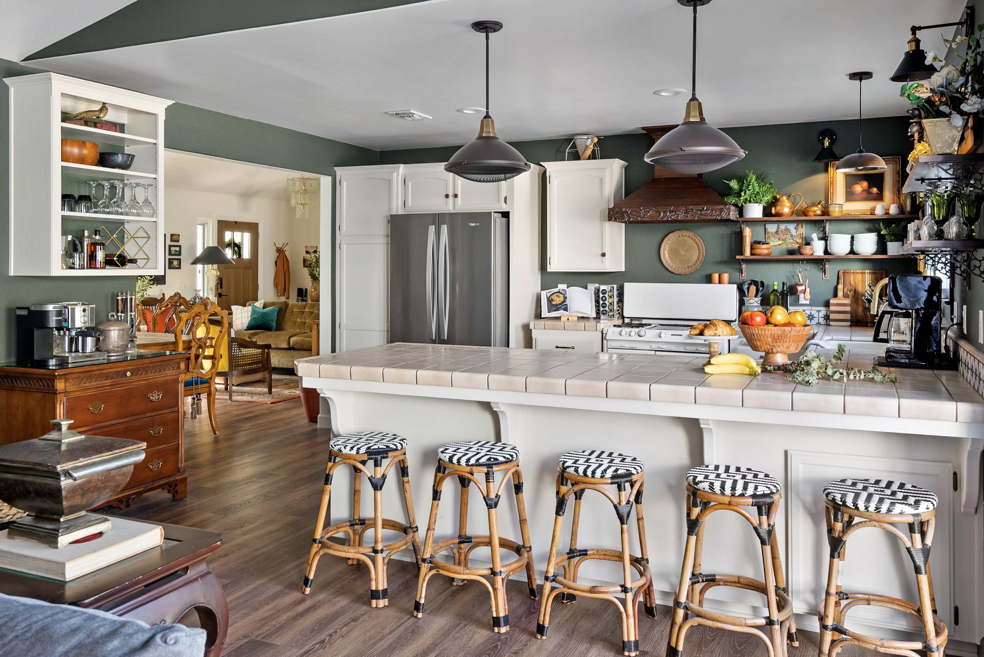 A modern kitchen features a central island with striped stools, stainless steel appliances, and open shelving, connected to a cozy living area in the background.