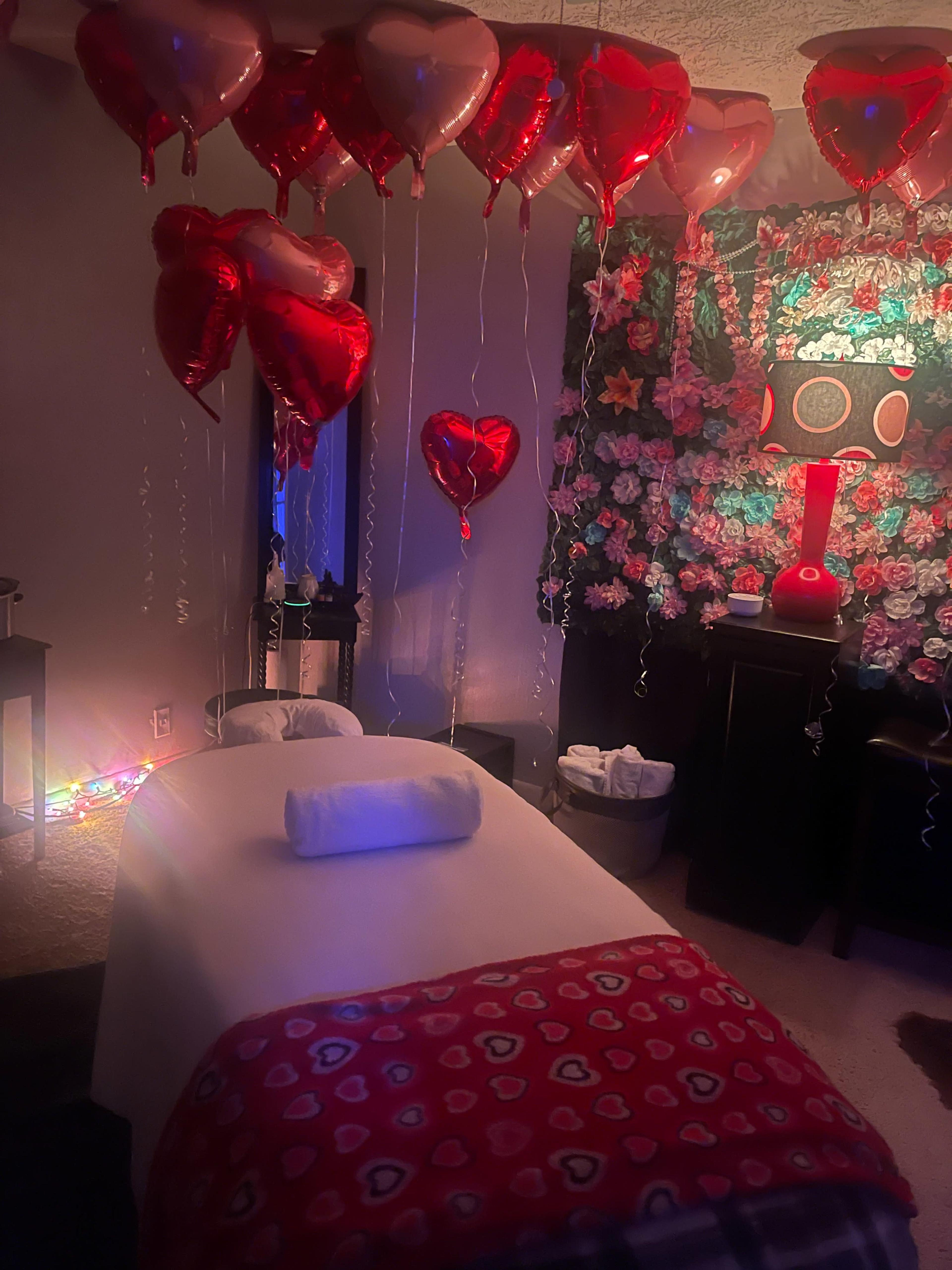 The room is decorated with heart-shaped balloons and floral wall decor, featuring a massage table covered with a blanket and a rolled towel.