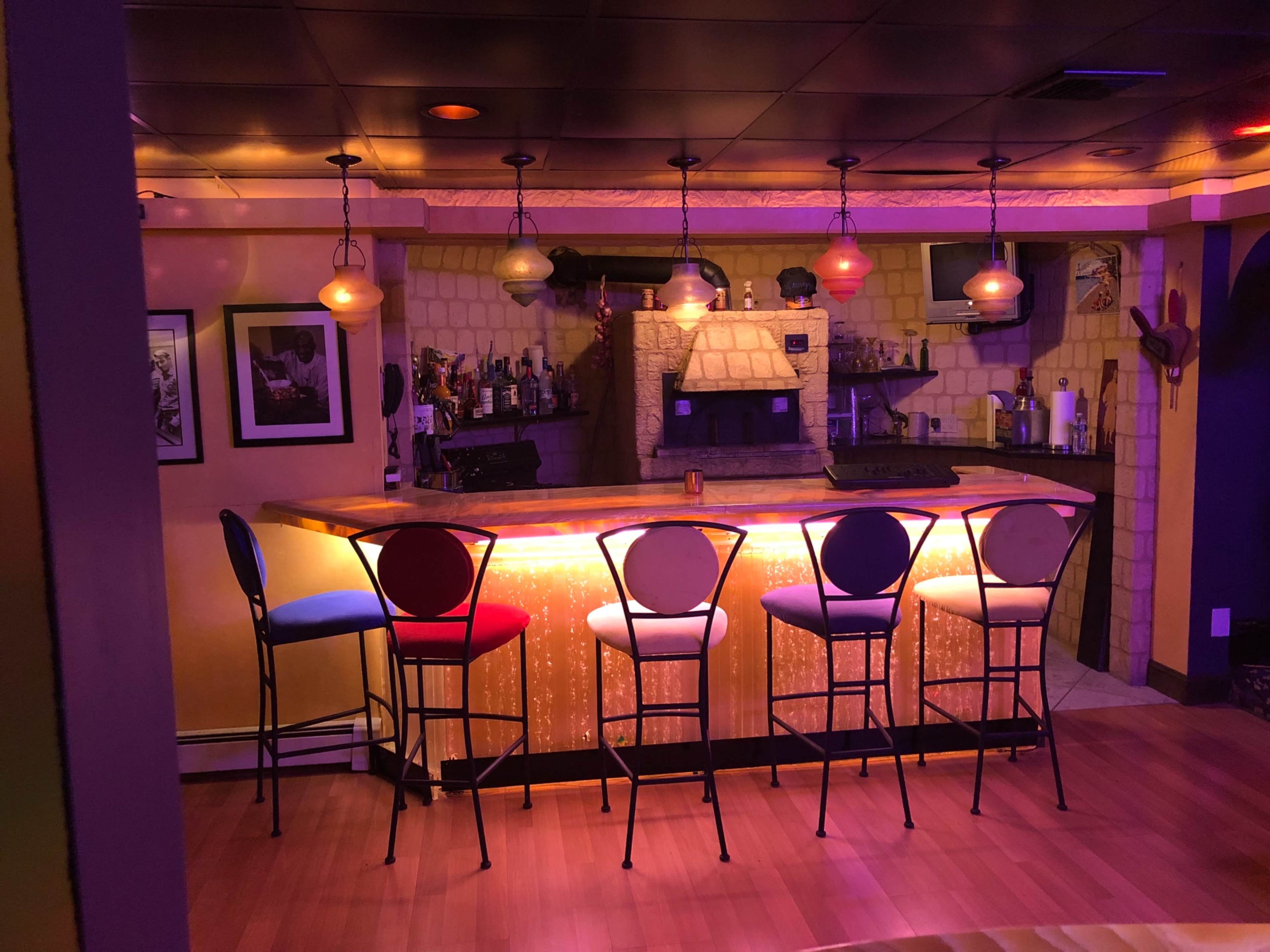 A well-lit home bar with colorful stools arranged around a counter, set against a backdrop of stone walls and shelves containing bottles.