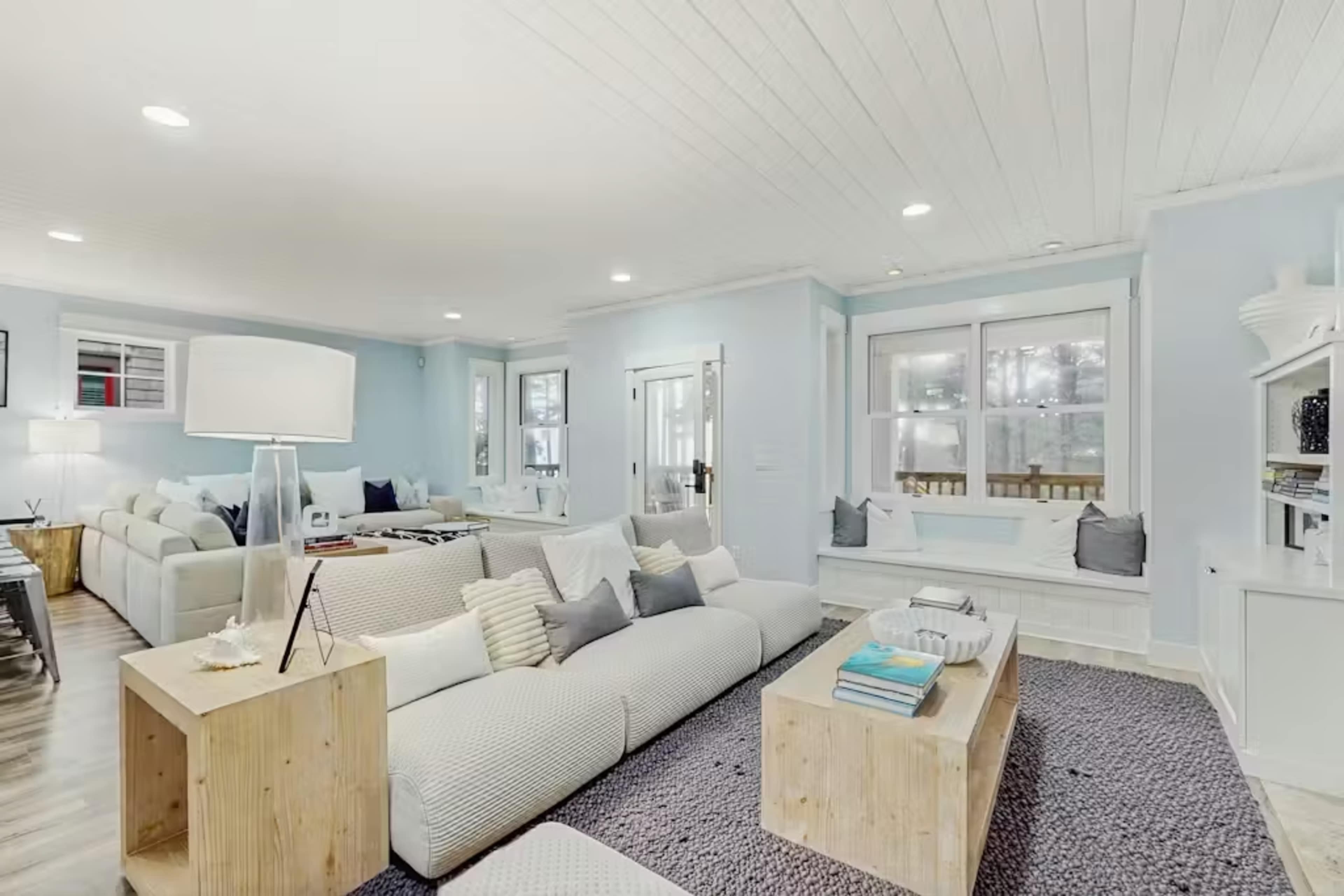 The image shows a spacious, light blue living room with a large sectional couch, a wooden coffee table, and large windows allowing natural light to enter.