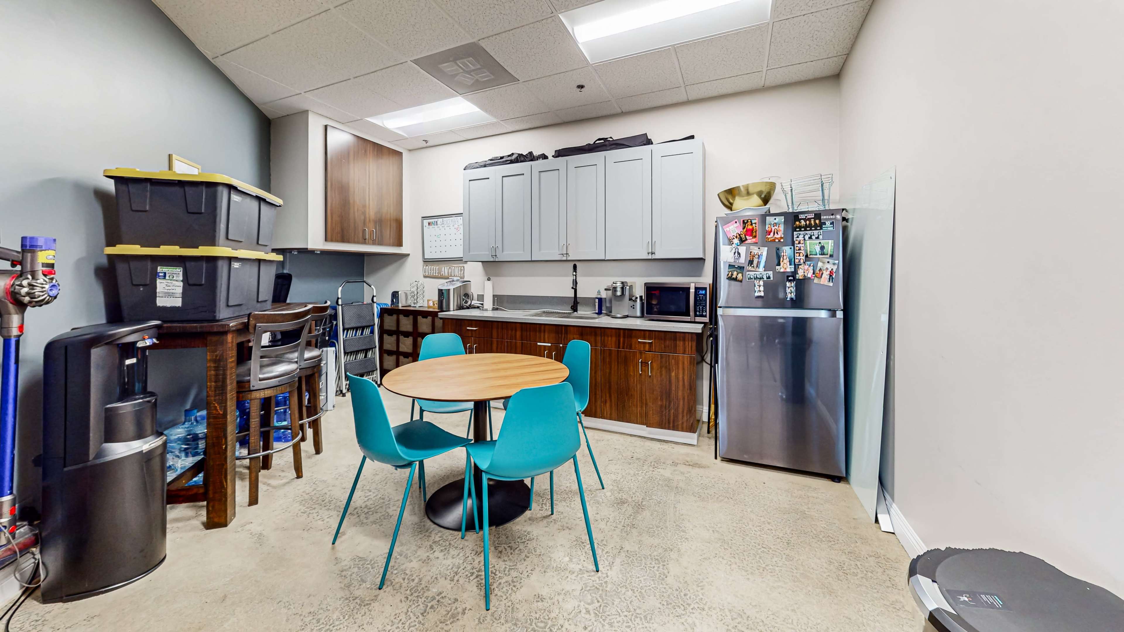 The image shows a small kitchenette with a round table and four blue chairs, alongside cabinets, a refrigerator, and various kitchen appliances.