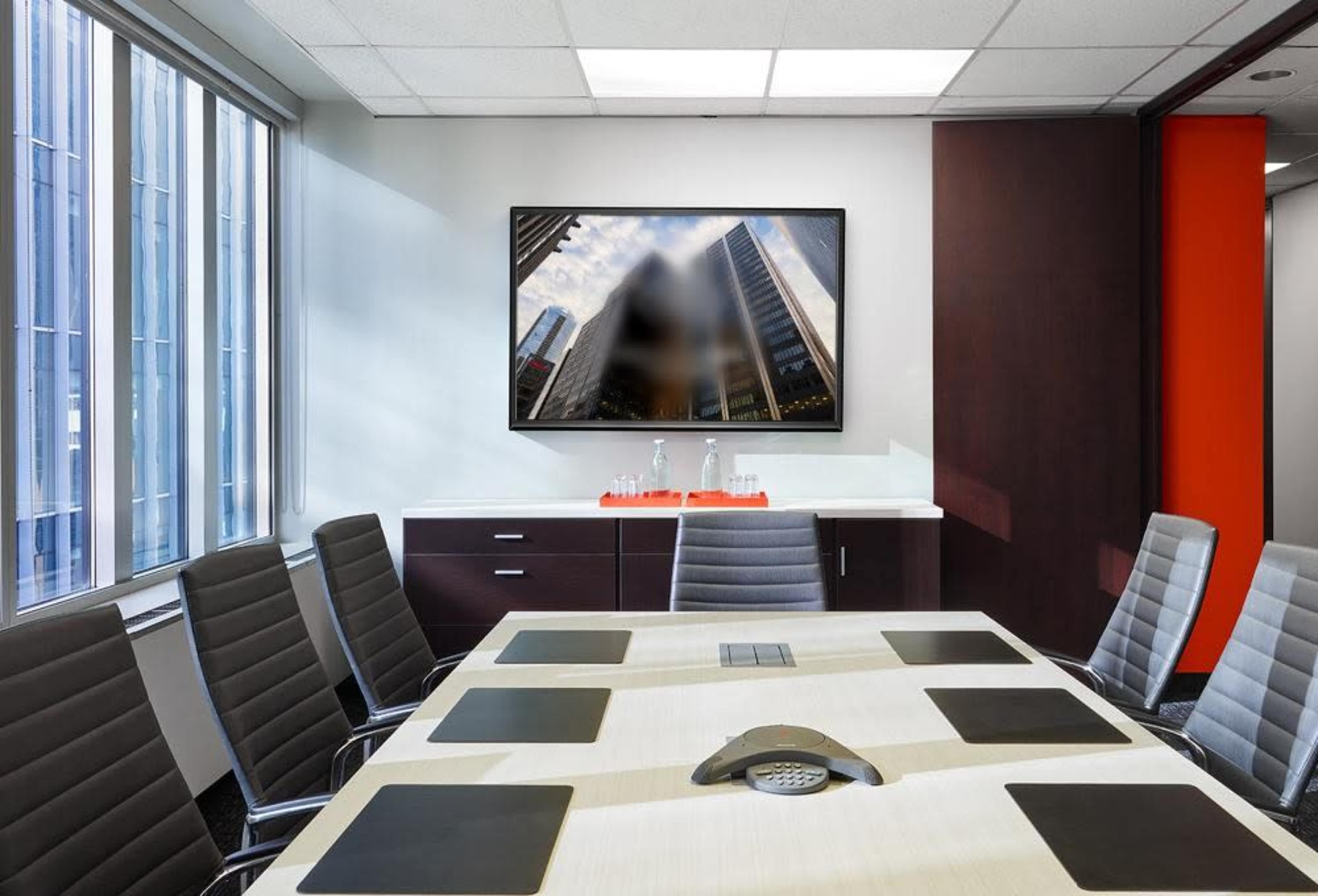 A boardroom features a large table surrounded by chairs and a wall-mounted television displaying a cityscape.
