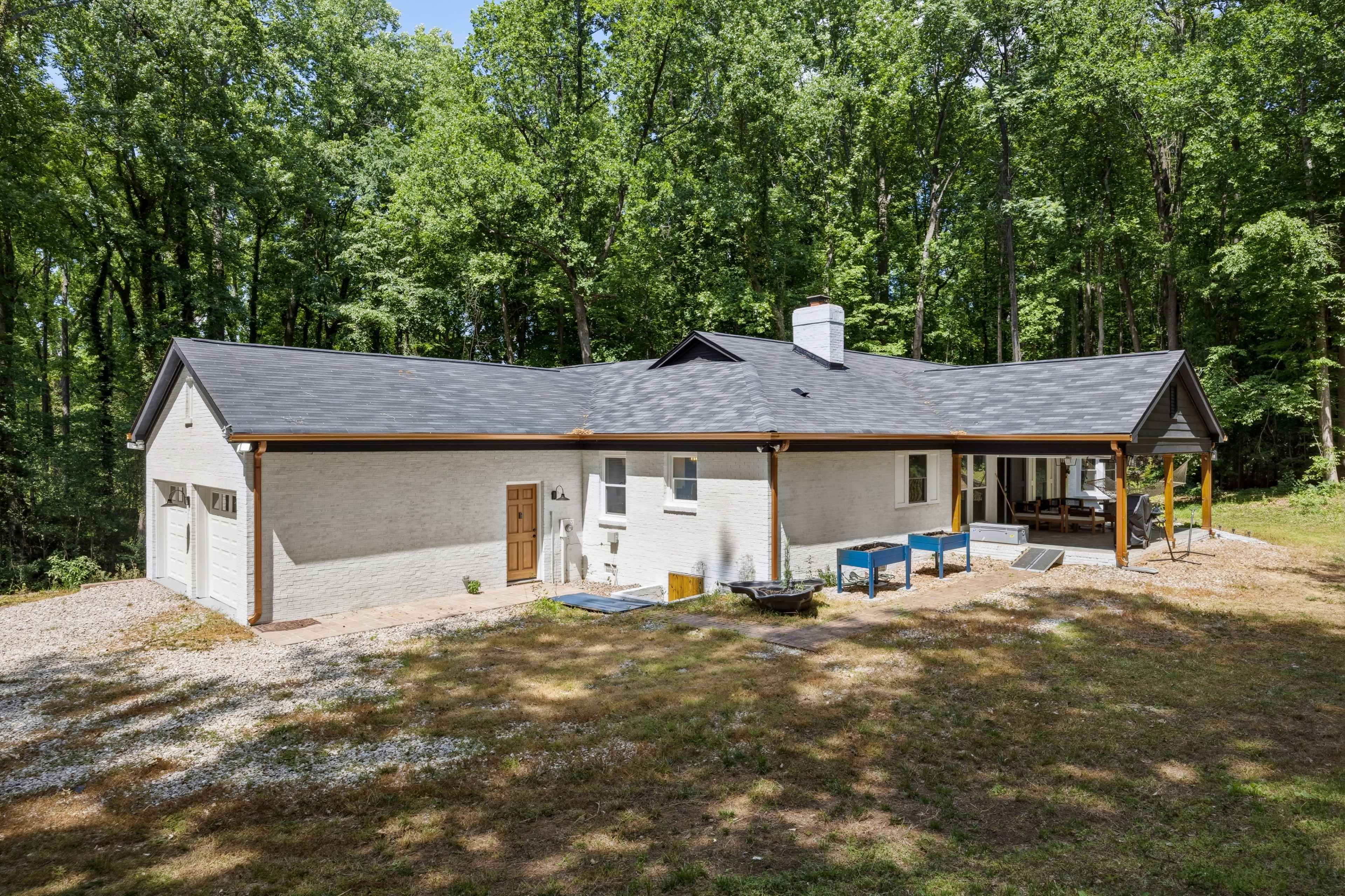 A modern single-story home with a white exterior and wooden accents is surrounded by dense green trees and a gravel yard.
