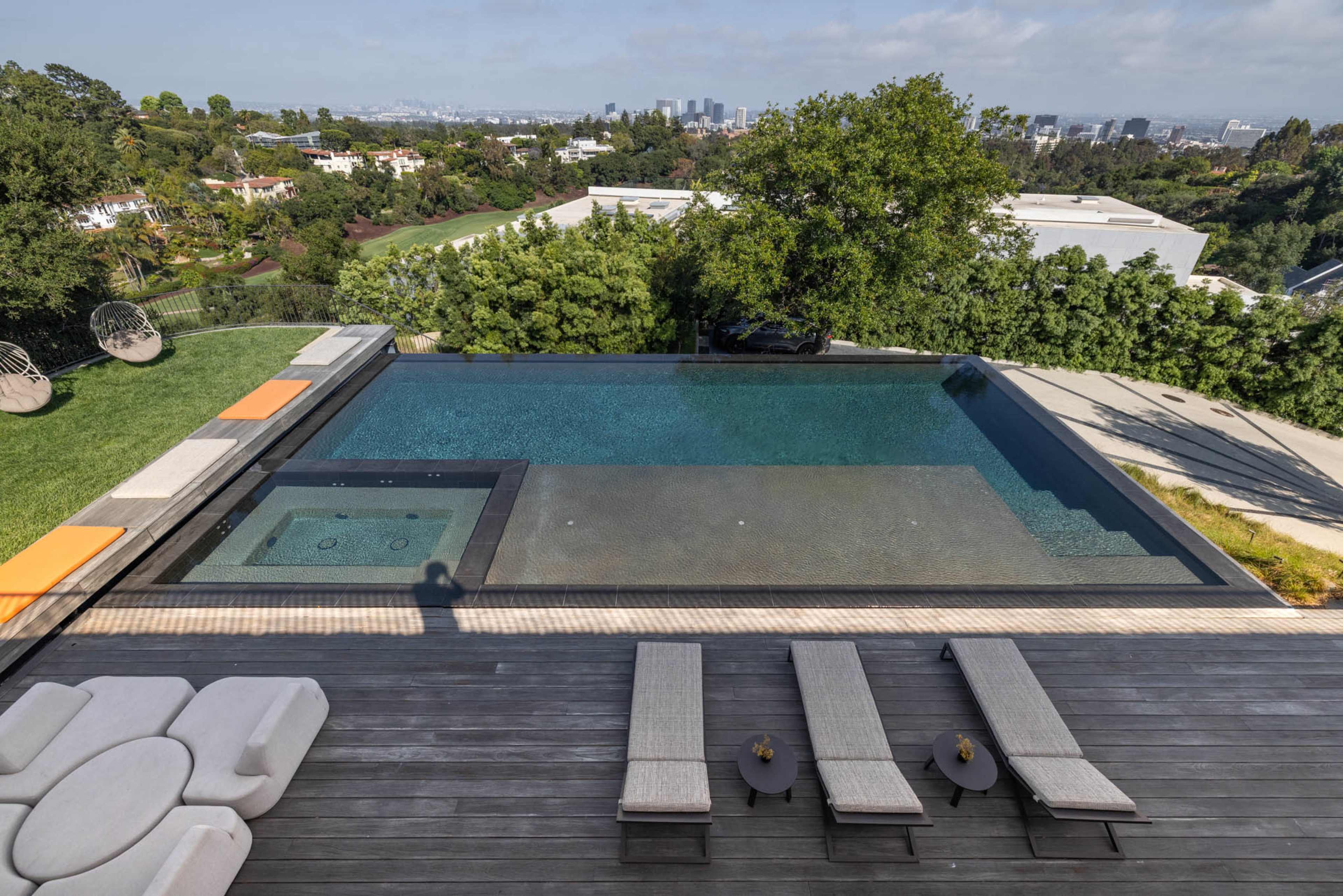The image shows a modern infinity pool with a hot tub overlooking a cityscape, surrounded by a wooden deck and lounge chairs.