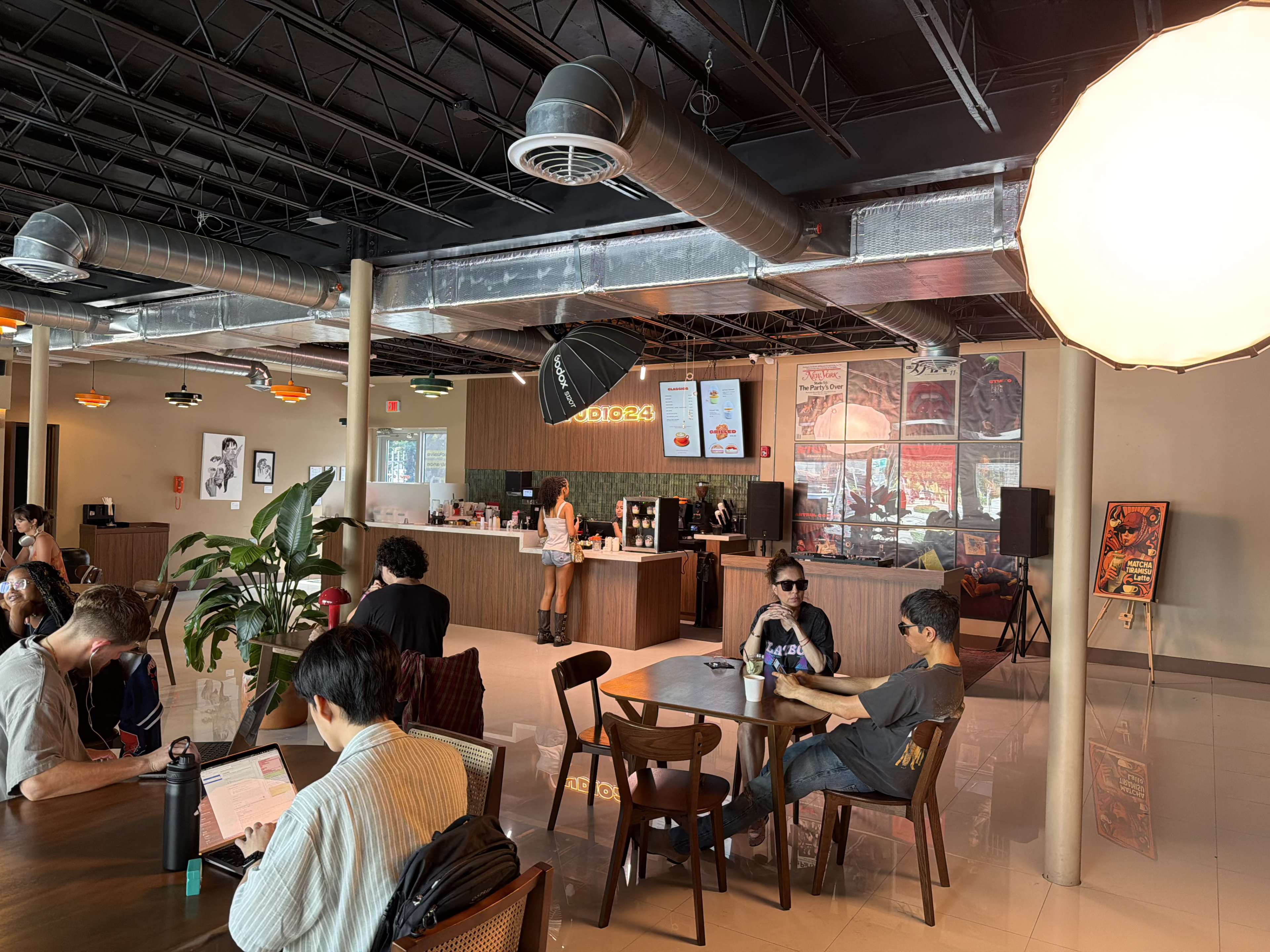 The image shows a modern cafe interior with patrons seated at wooden tables, a service counter in the background, and artwork on the walls.