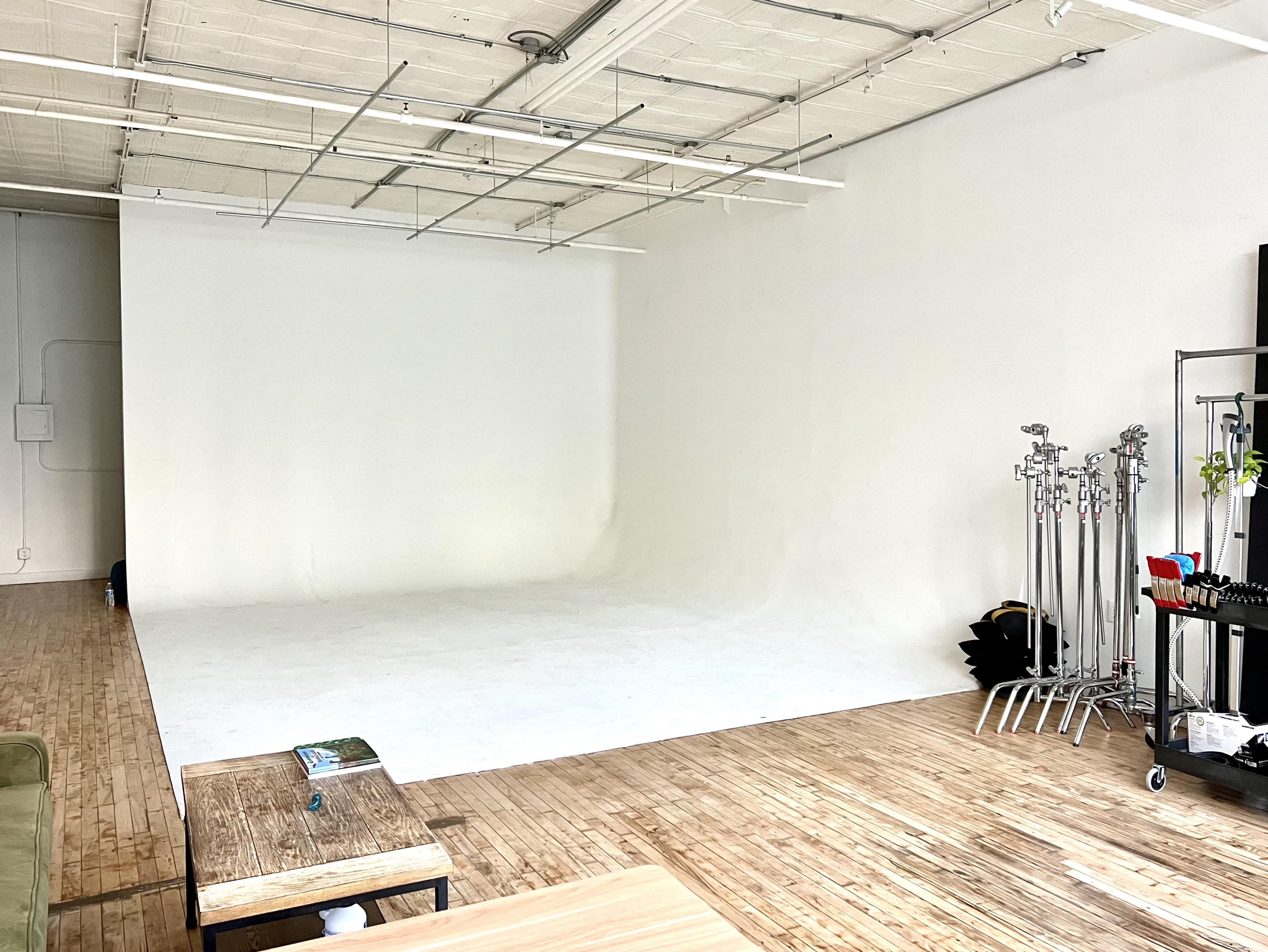 A spacious, empty photography studio with a white backdrop and wooden flooring, along with a coffee table and equipment stands on one side.
