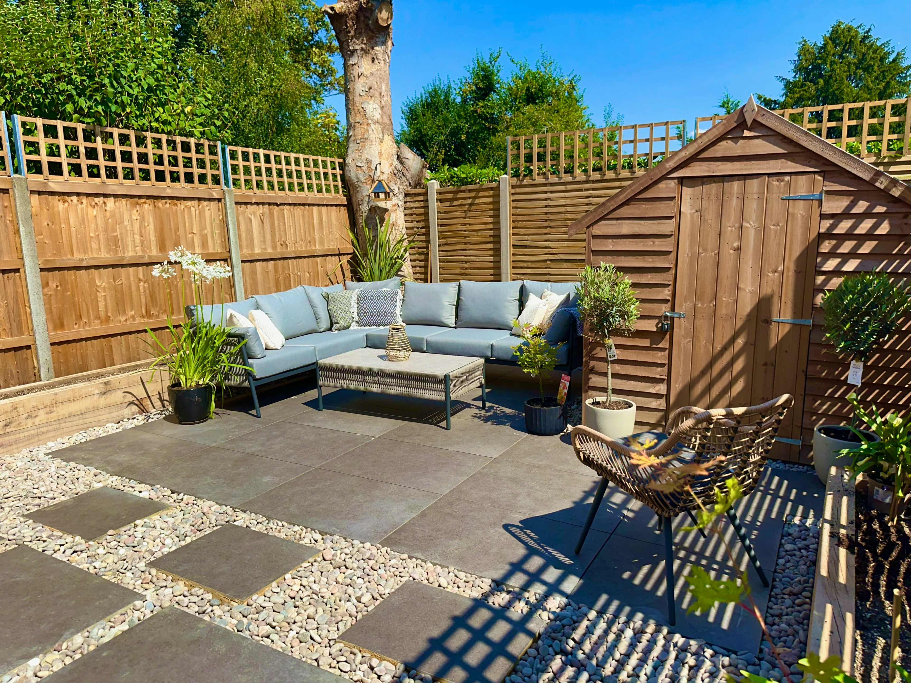 The image shows a landscaped outdoor space with a sectional sofa, a small table, potted plants, a garden shed, and decorative stone pathways.